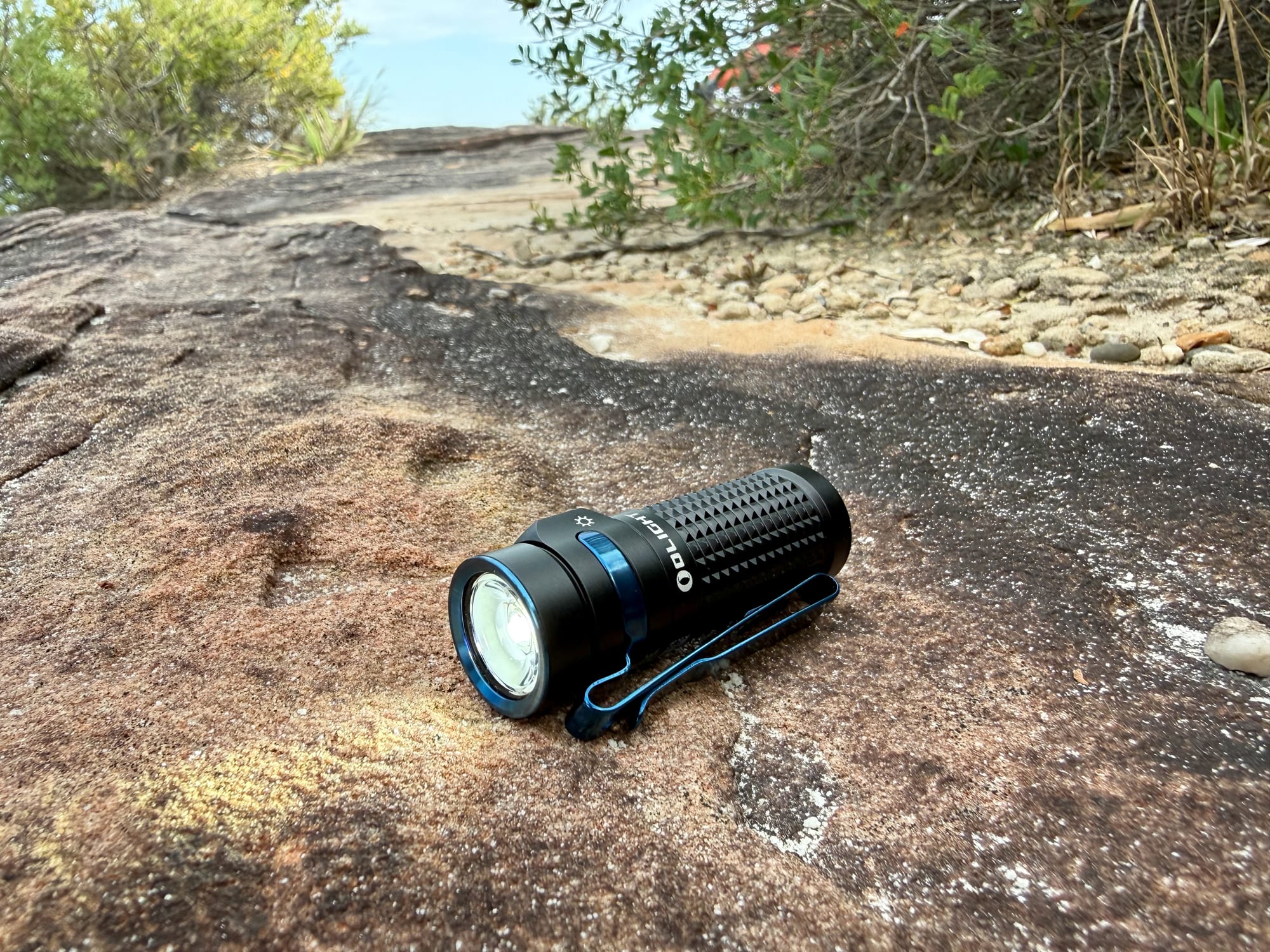 The Olight Baton 4 on its own, switched on, lying on a sandstone rock with bush behind