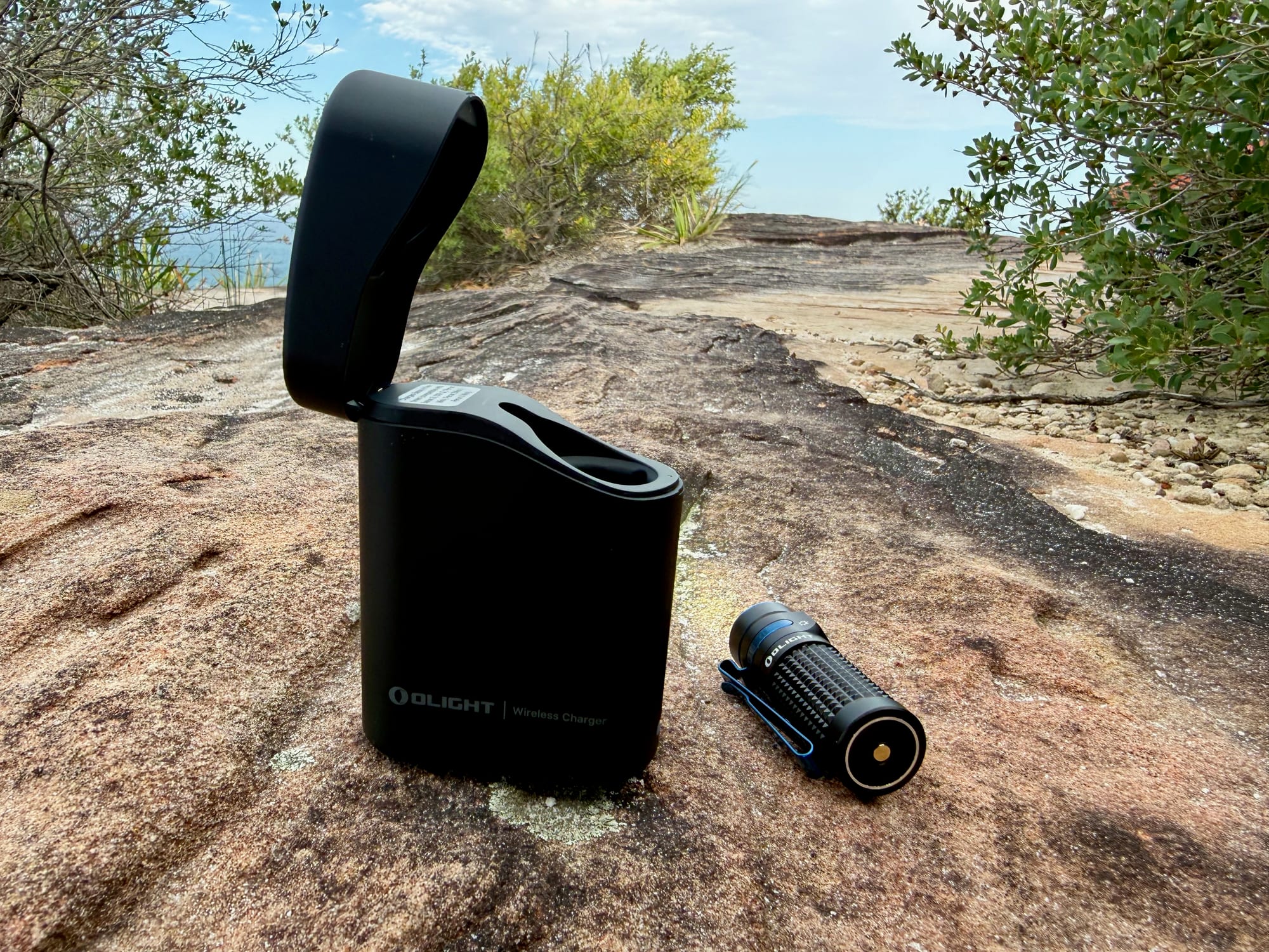 The Olight Baton 4 Premium Edition charging case with the lid open and the torch beside it, on a rock