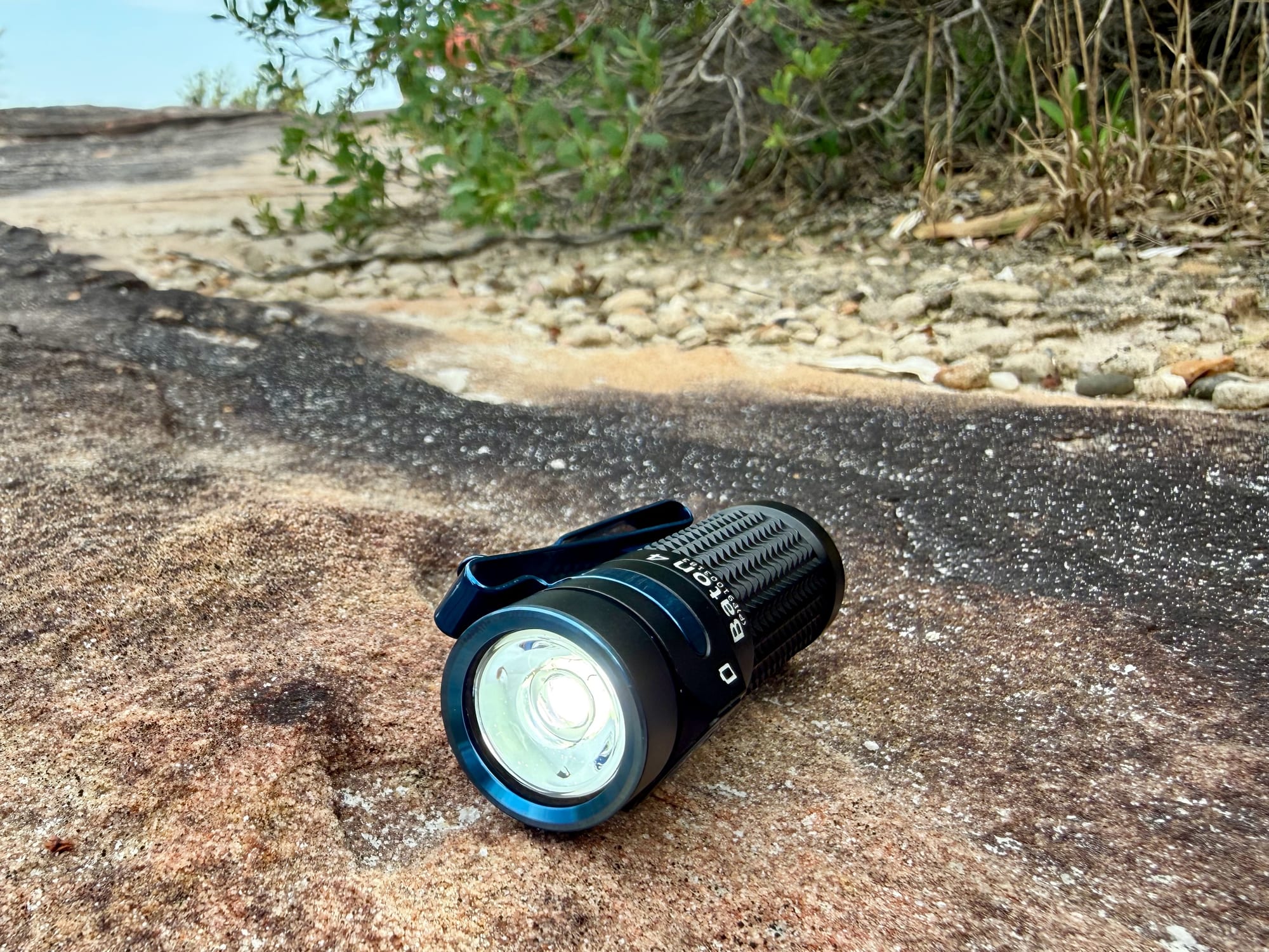 The Olight Baton 4 switched on, lying on a sandstone rock with bush behind