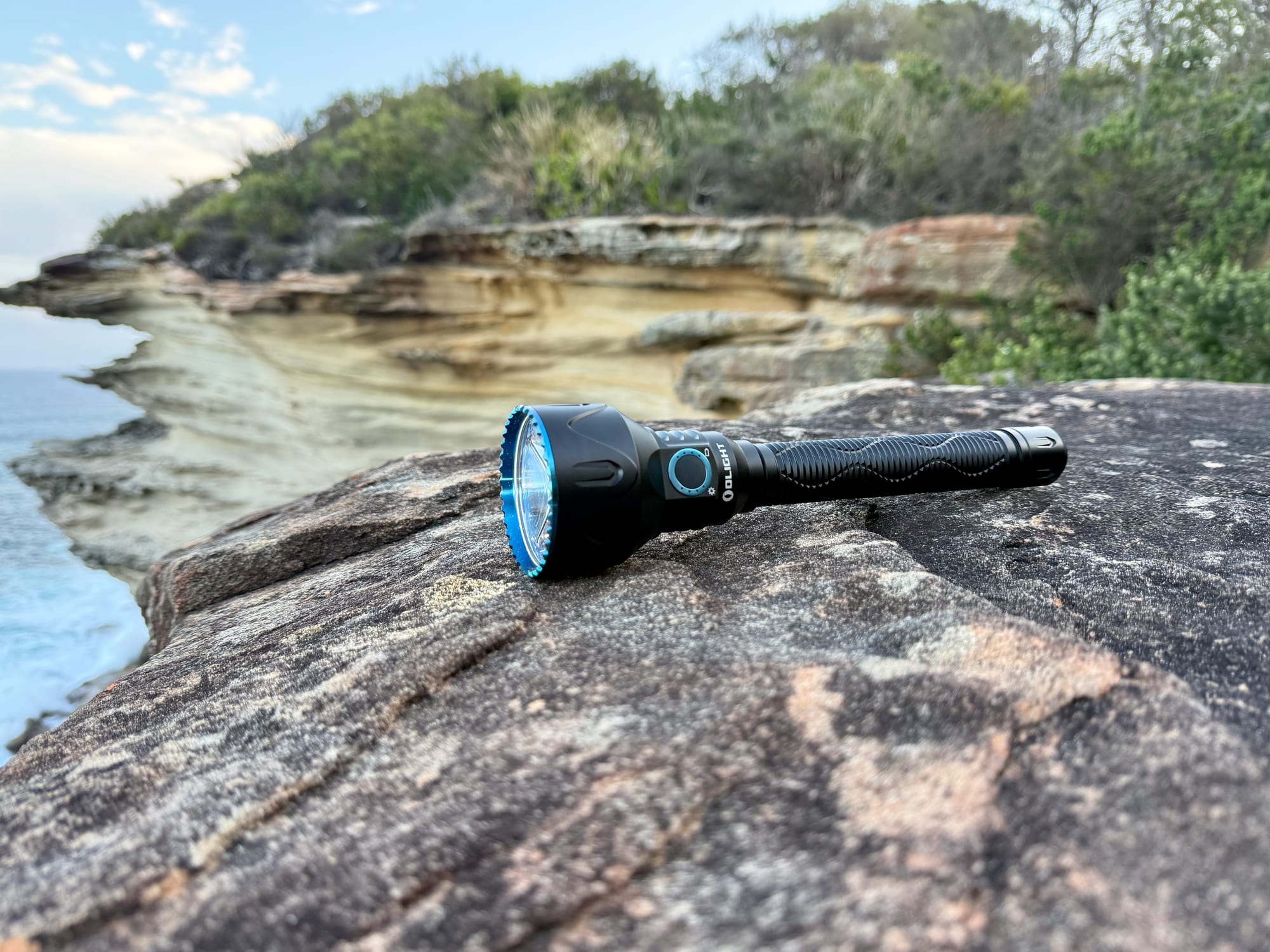 The Olight Javelot Pro 2 lying on a sandstone rock above the sea, with cliffs and bush behind