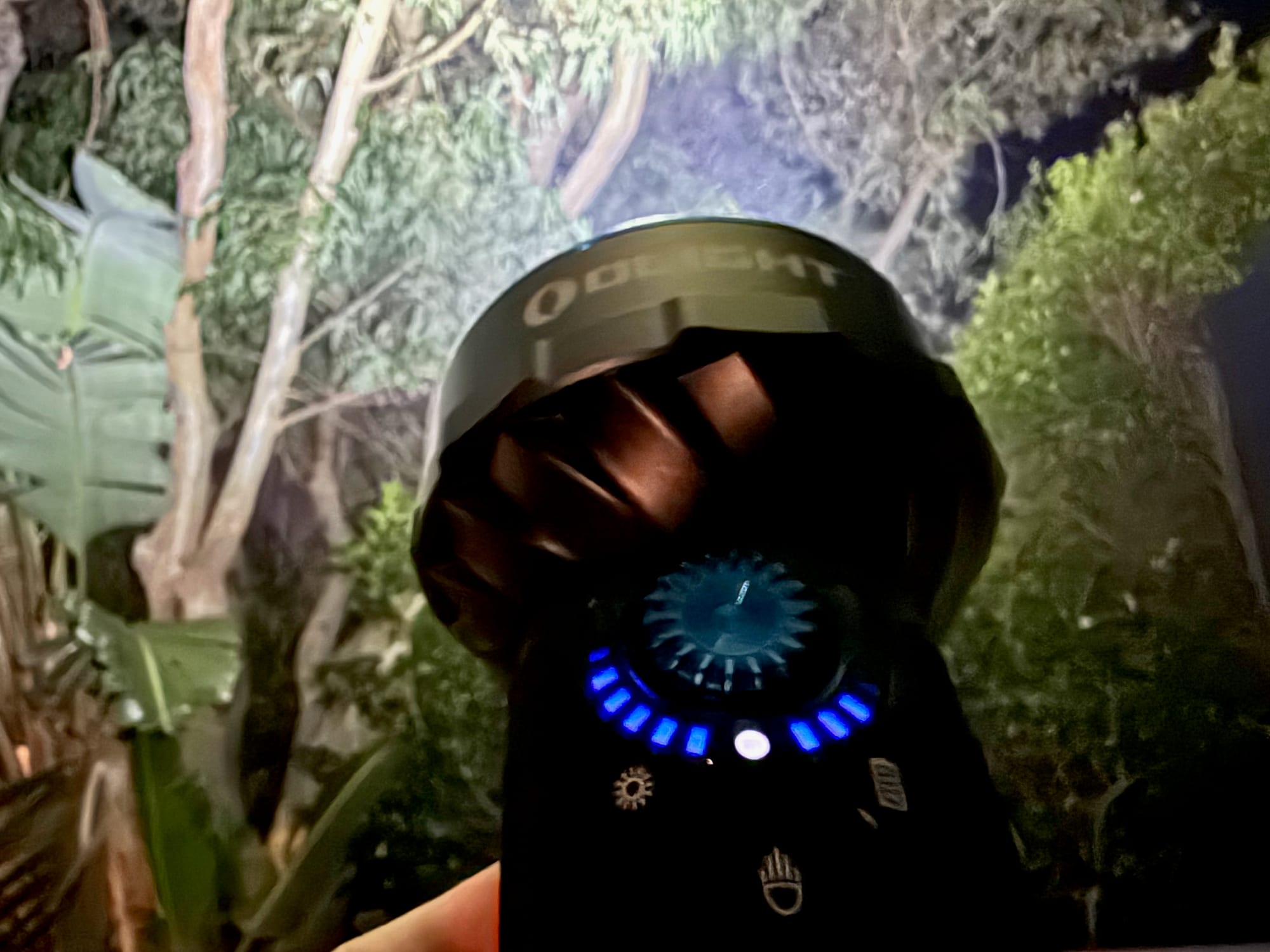 The Olight Marauder Mini held up at night, its flood lighting up the trees and the blue brightness ring around the dial lit