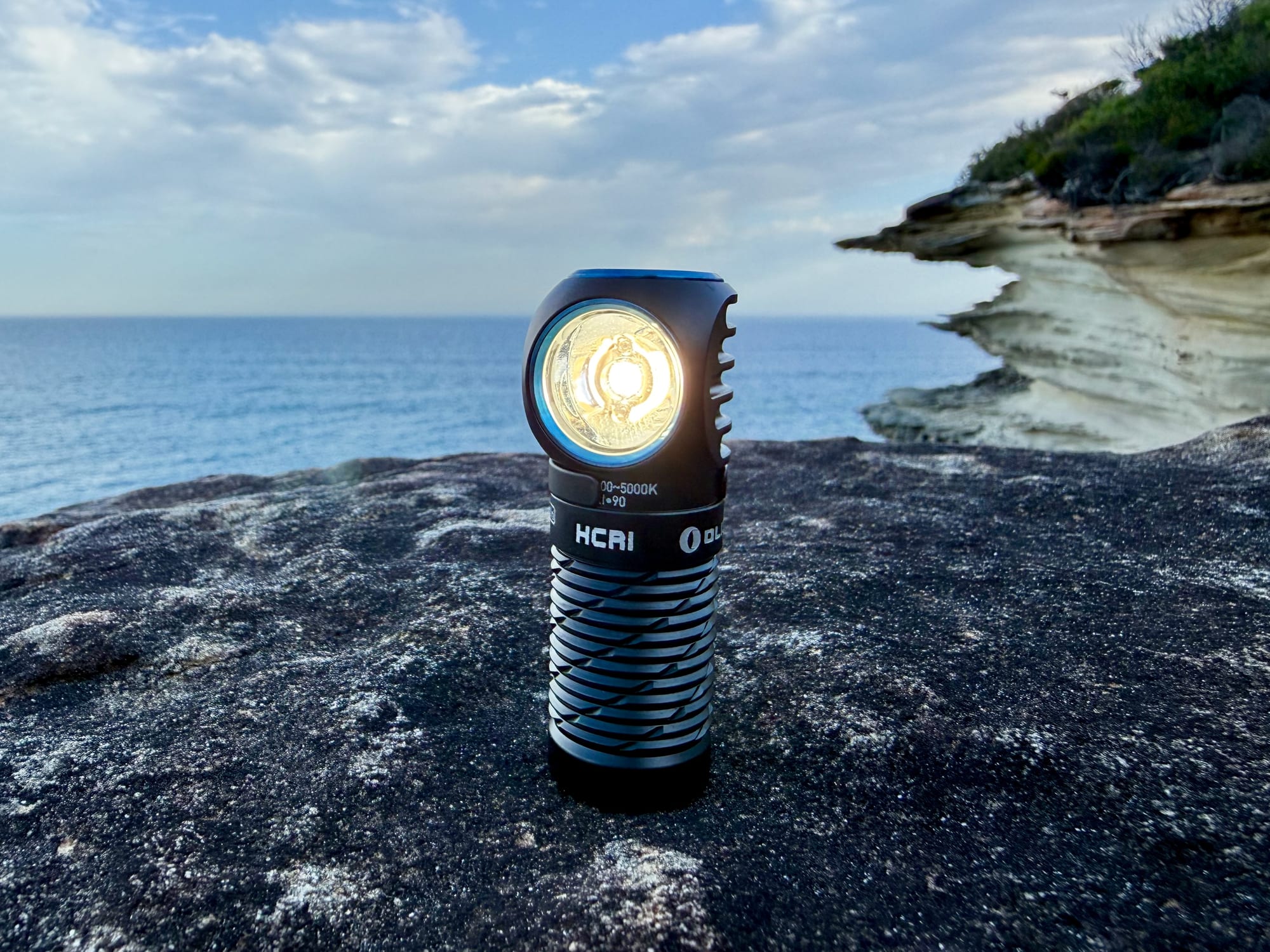 The Olight Perun 3 Mini standing on a rock with its neutral white light on, HCRI on the body, sea and cliffs behind