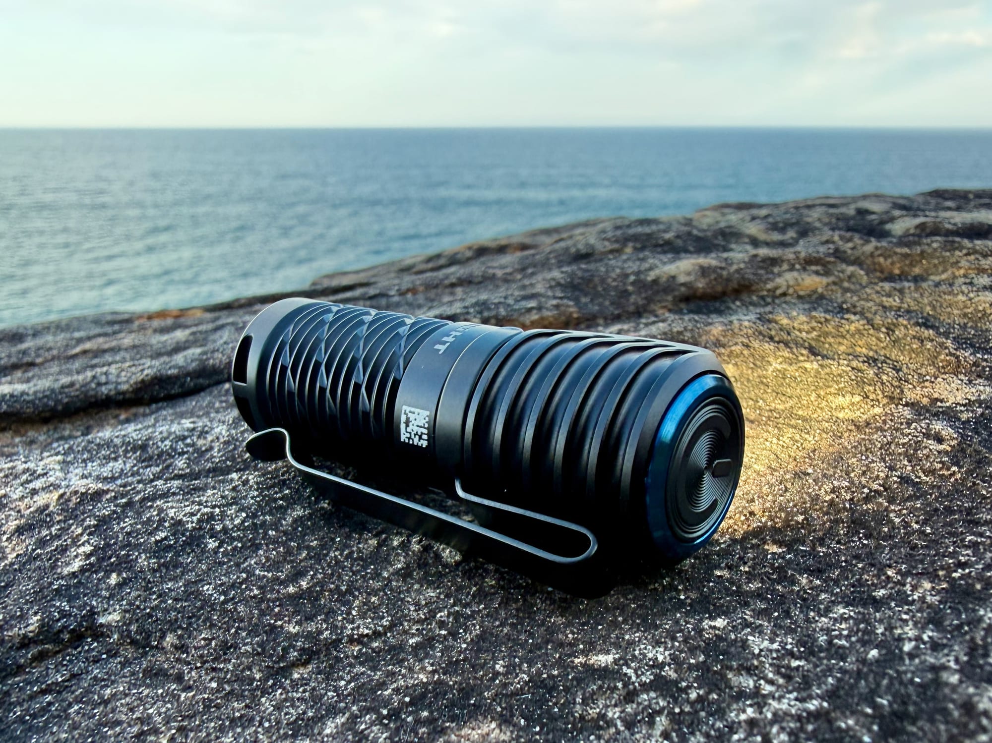 The Olight Perun 3 Mini lying on a rock by the sea, showing the pocket clip and the power button on top of the head