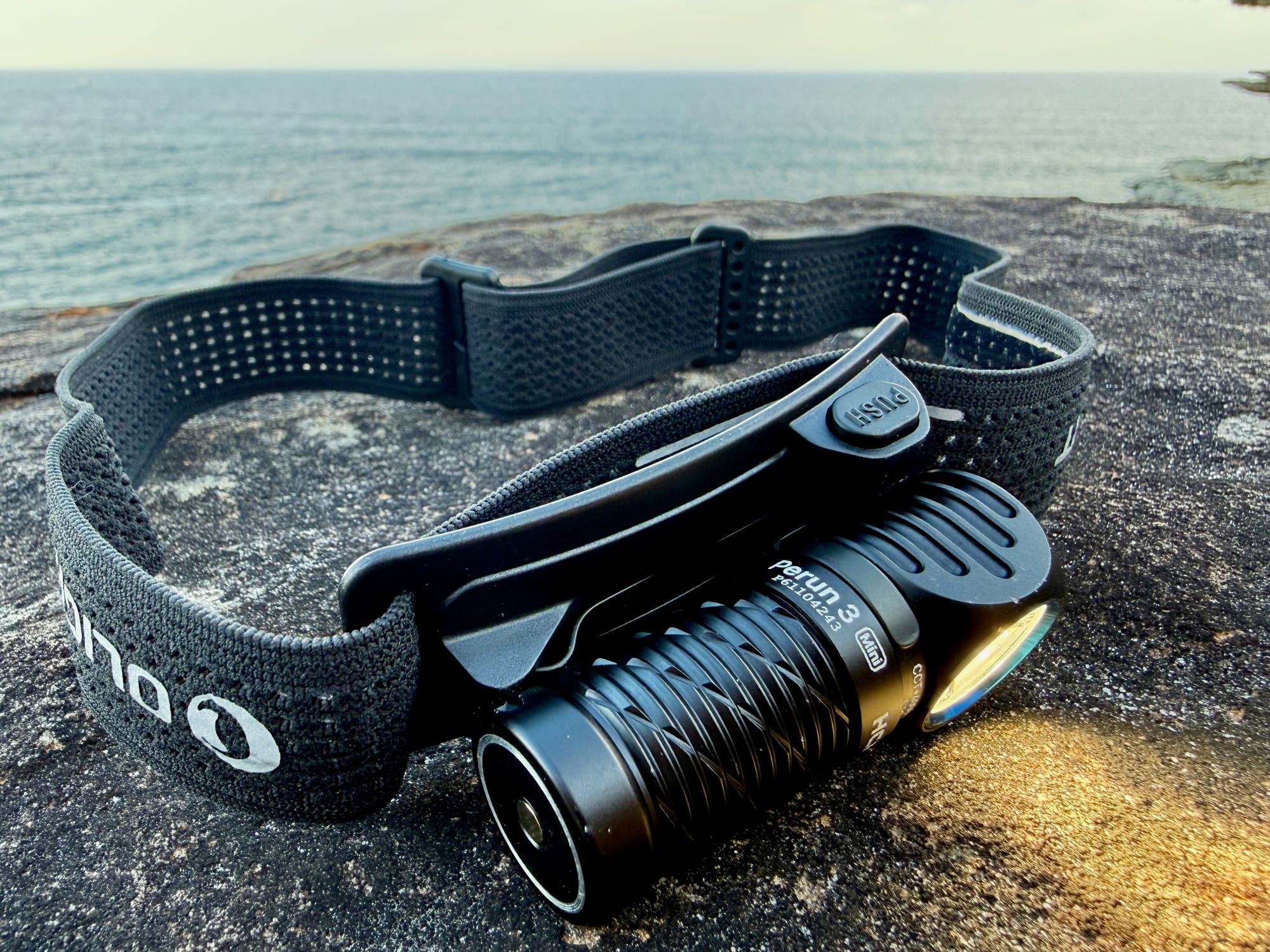 The Perun 3 Mini switched on in the plastic bracket on its mesh headband, with the PUSH release tab, by the sea