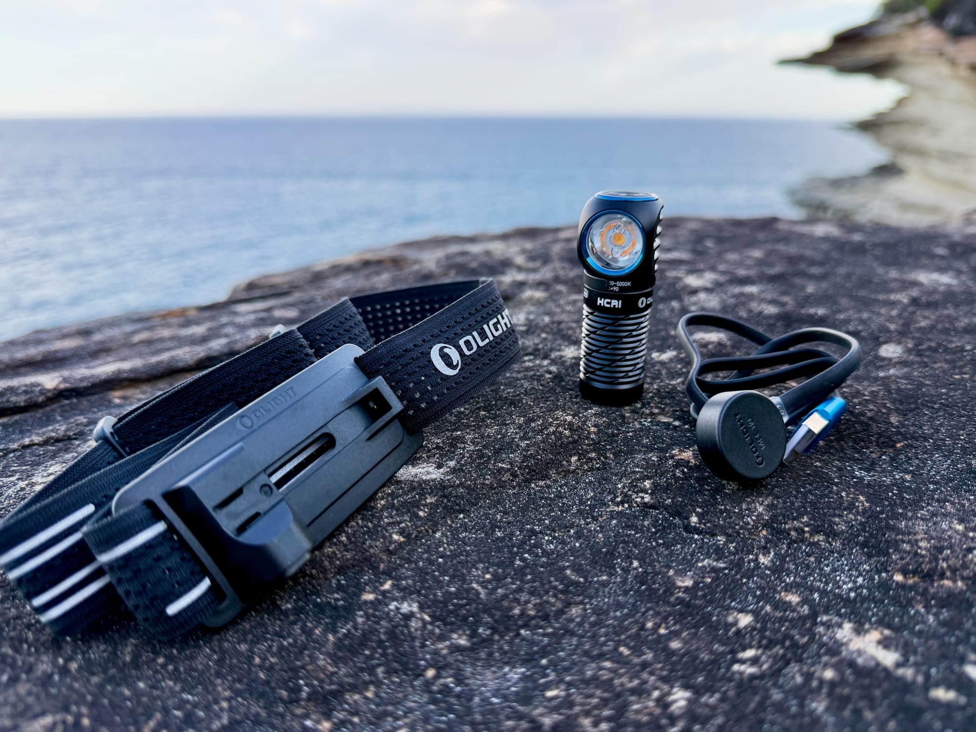 The Olight Perun 3 Mini with its mesh headband and magnetic charging cable laid out on a rock by the sea