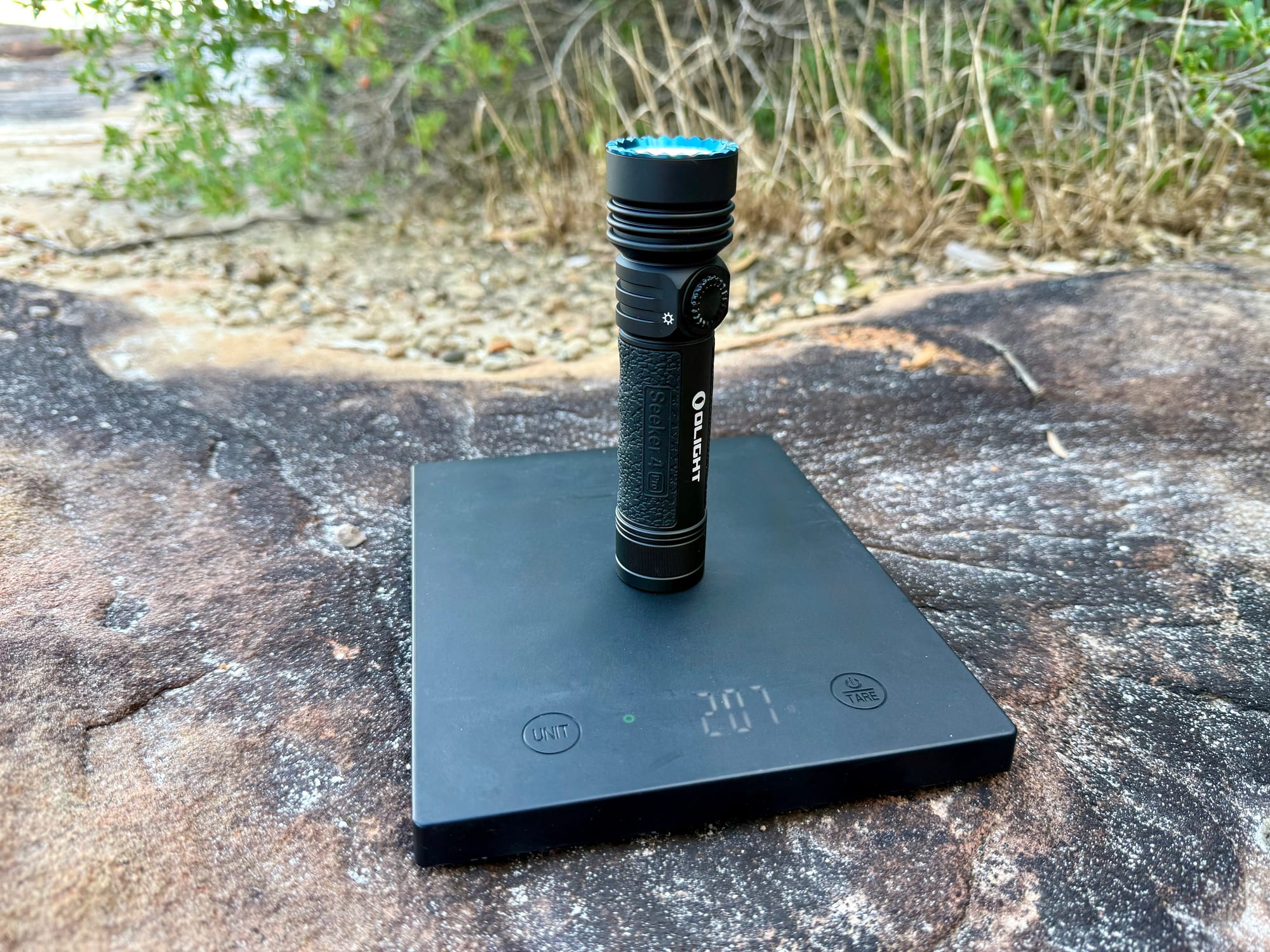 The Olight Seeker 4 Pro standing on a kitchen scale showing 207 g, with bush behind