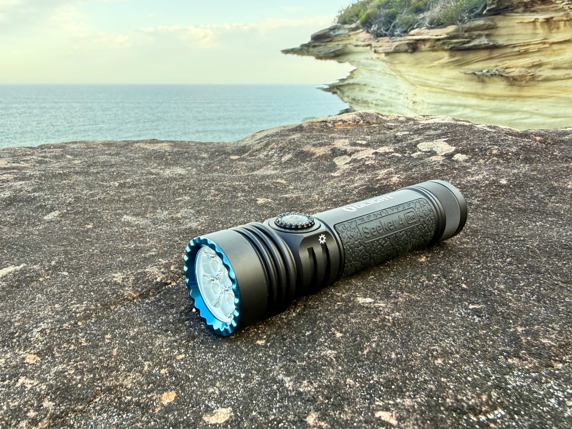 The Olight Seeker 4 Pro switched on, lying on a rock with the sea behind