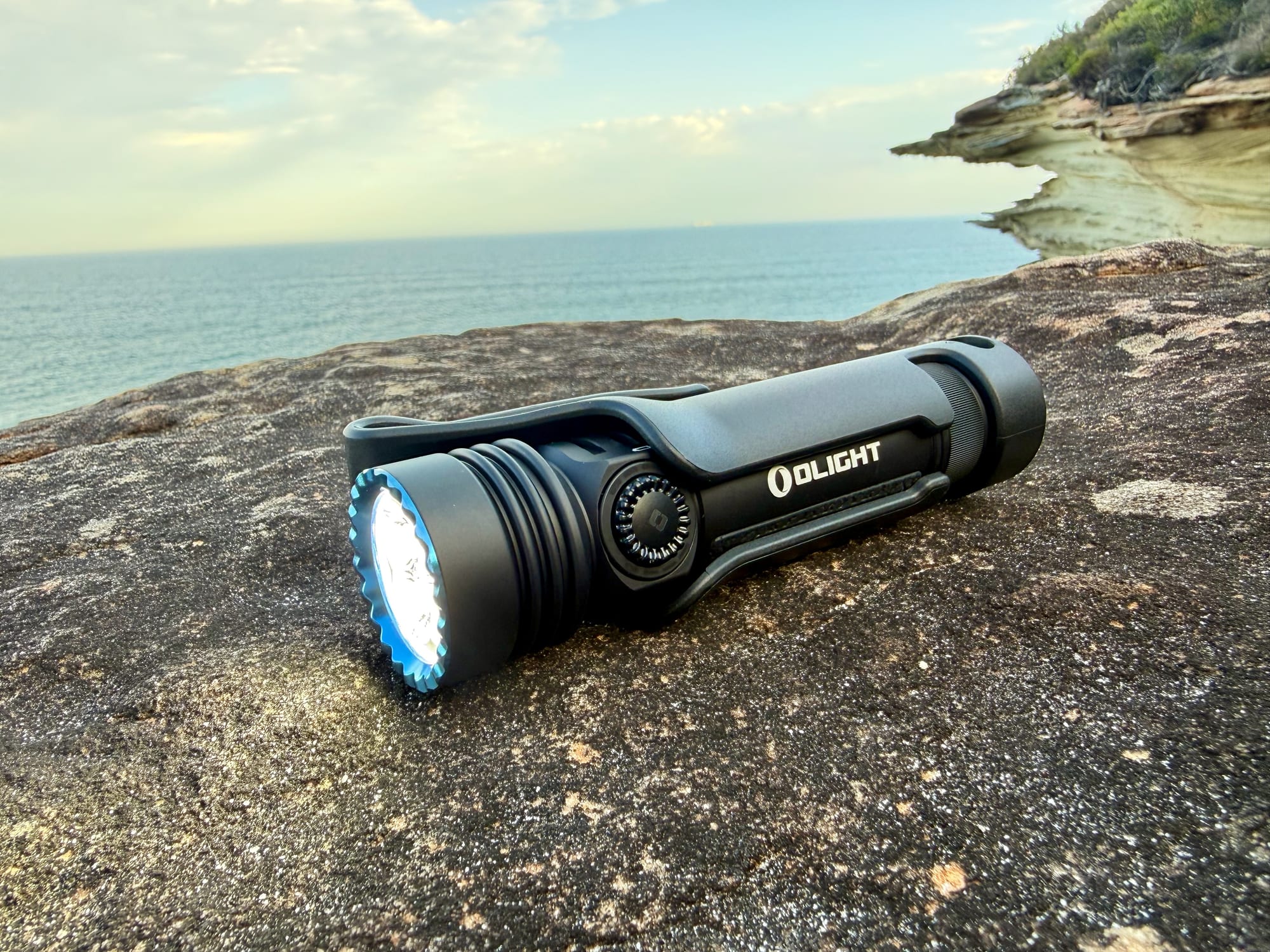 Olight Seeker 4 Pro torch switched on, lying on a rock above the sea with its pocket clip