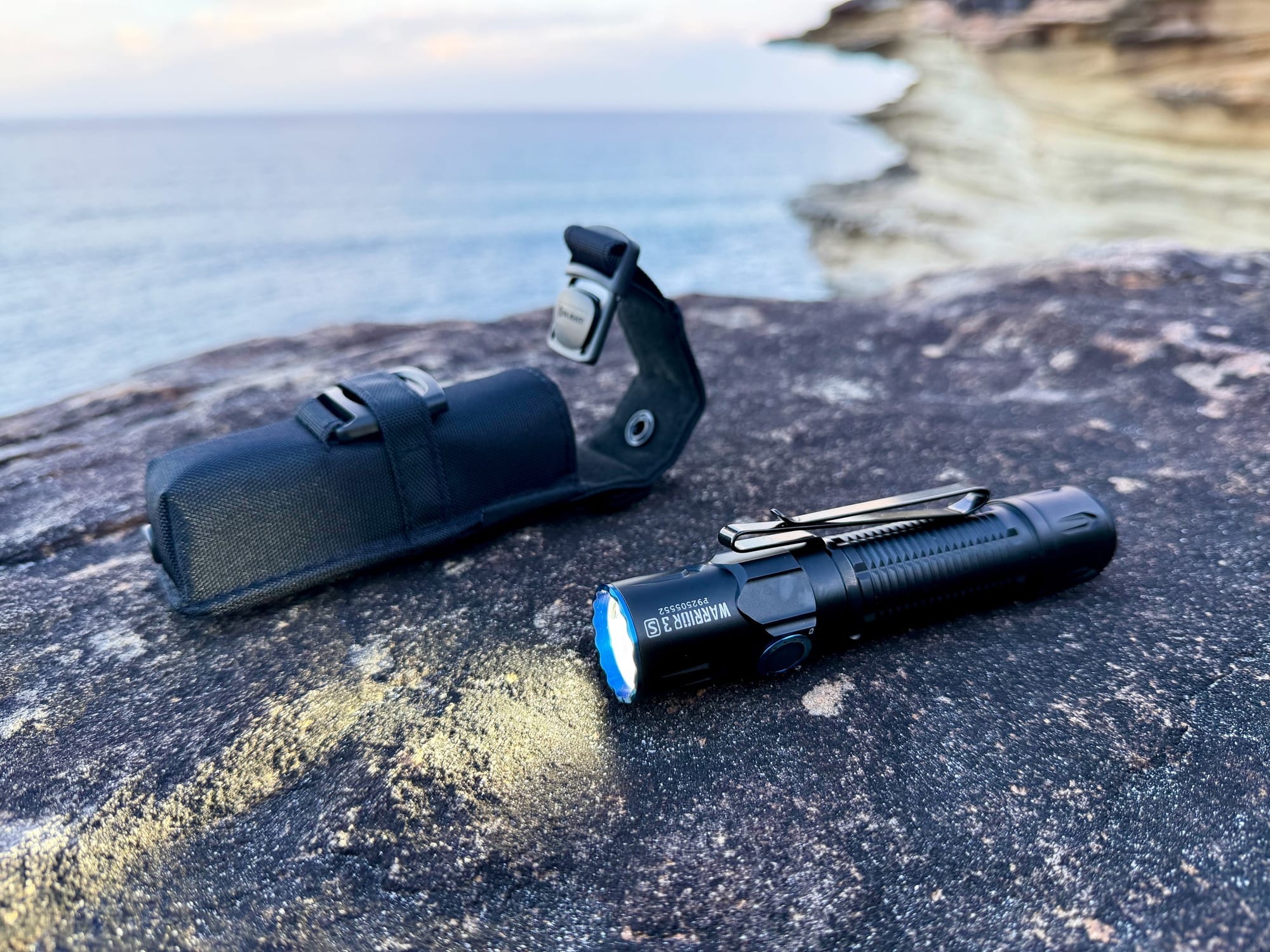 The Olight Warrior 3S next to its nylon holster on a sandstone rock above the sea