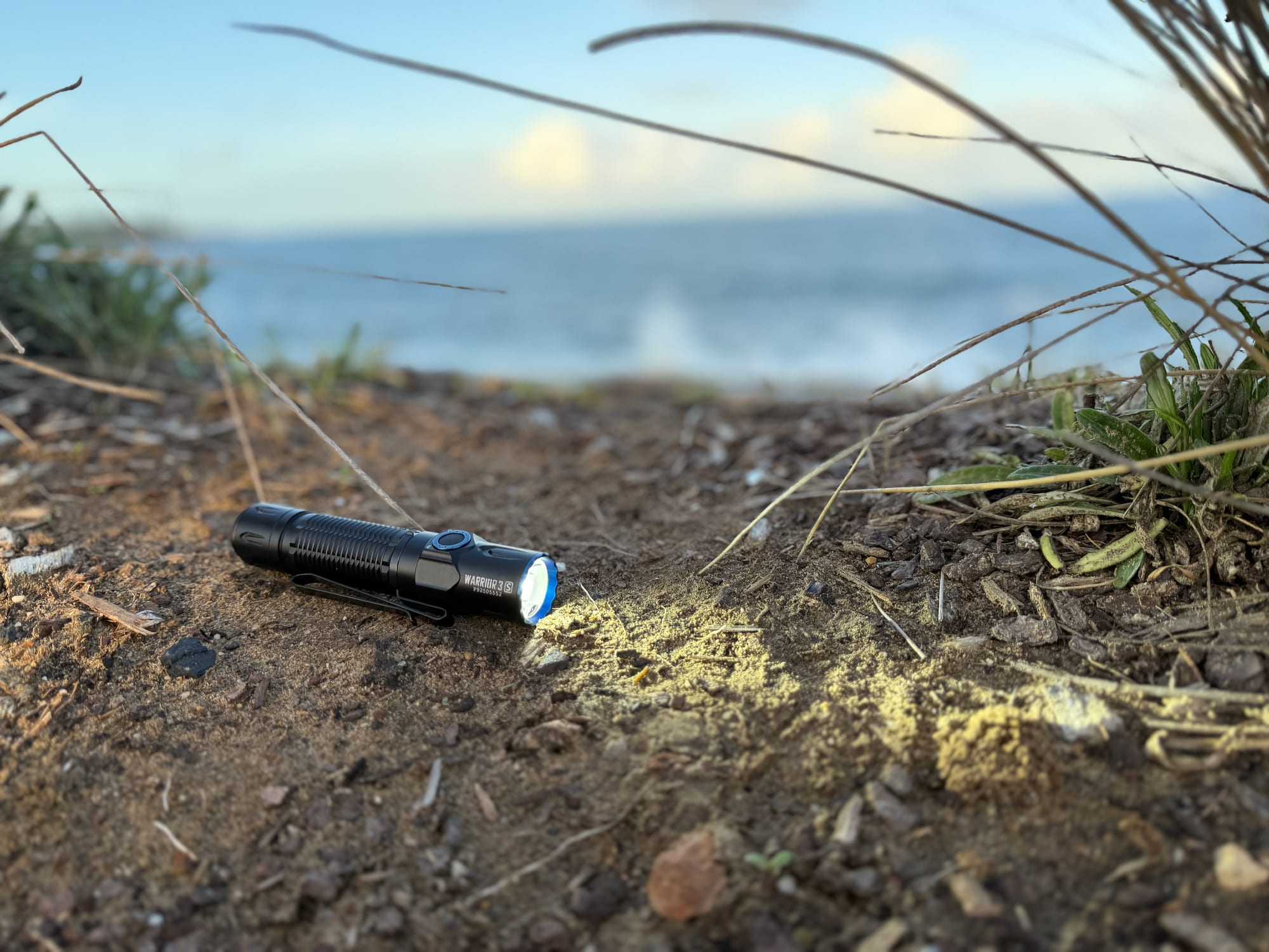 The Olight Warrior 3S switched on, its beam lighting up the ground on a headland above the sea