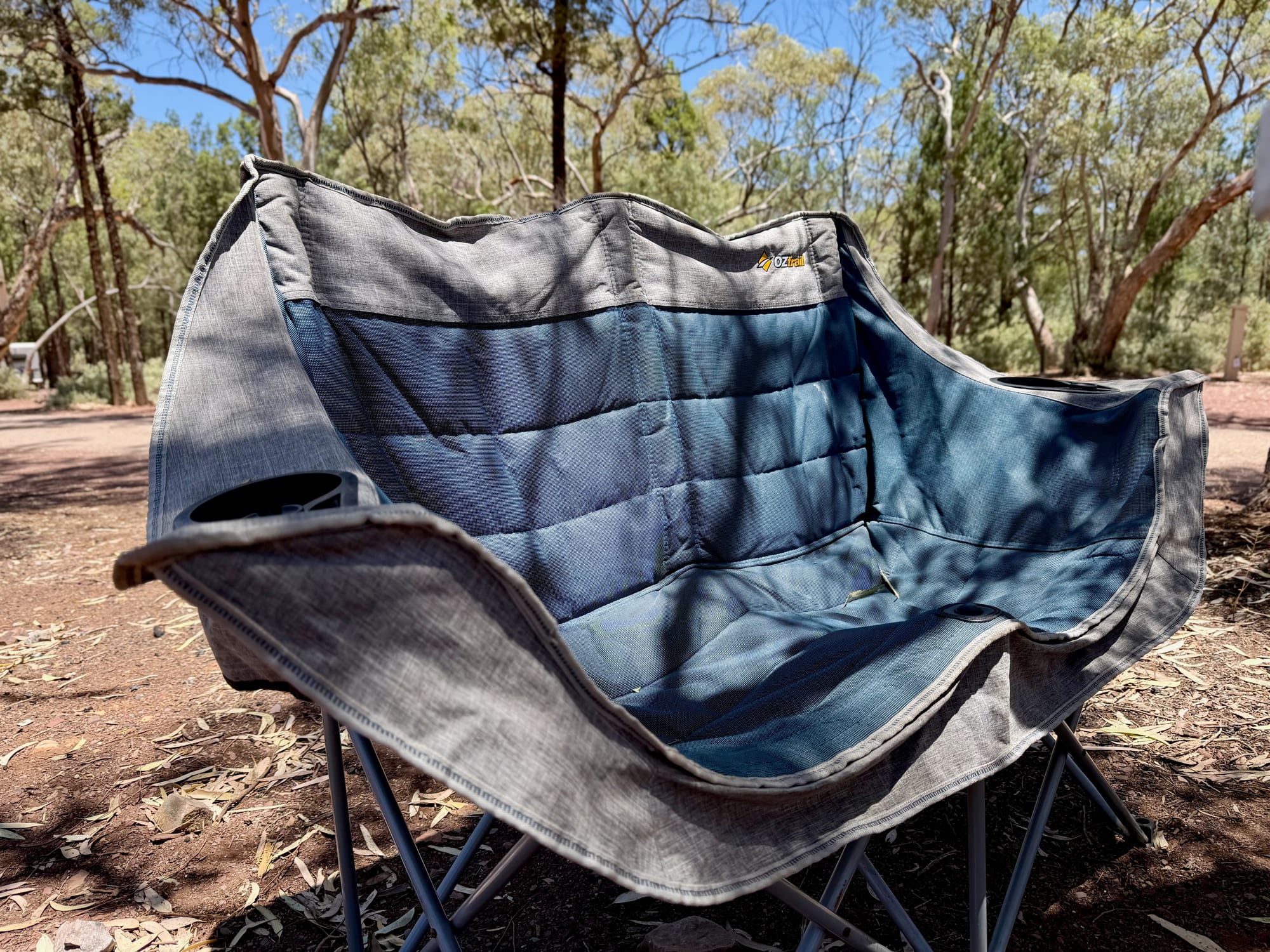 The OZtrail Galaxy 2 seater from the front, with its quilted blue padded seat, a cup holder in each arm and the middle support at the front edge