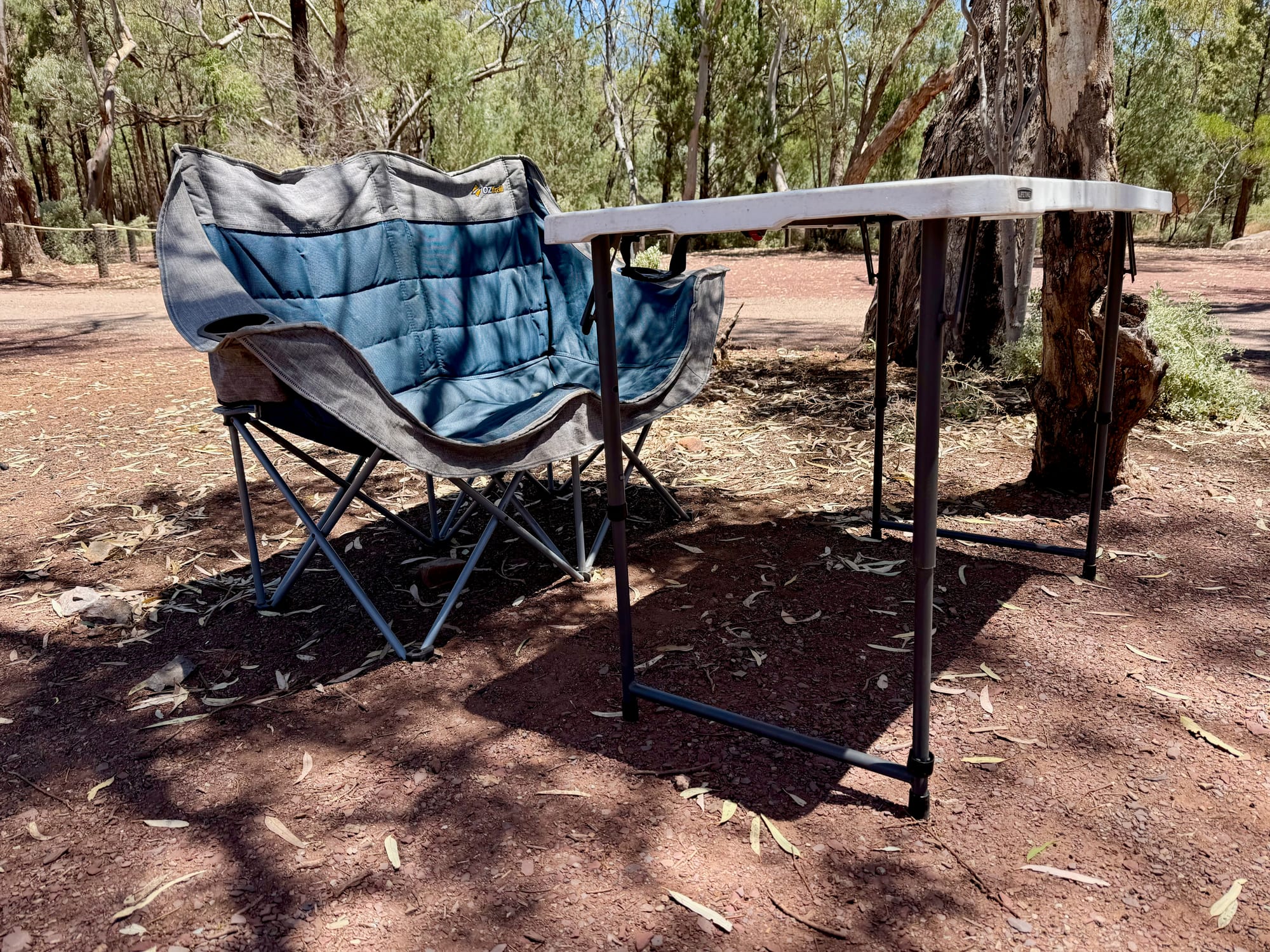 The OZtrail Galaxy 2 seater next to a folding table at a campsite among gum trees, with the table top well above the seat