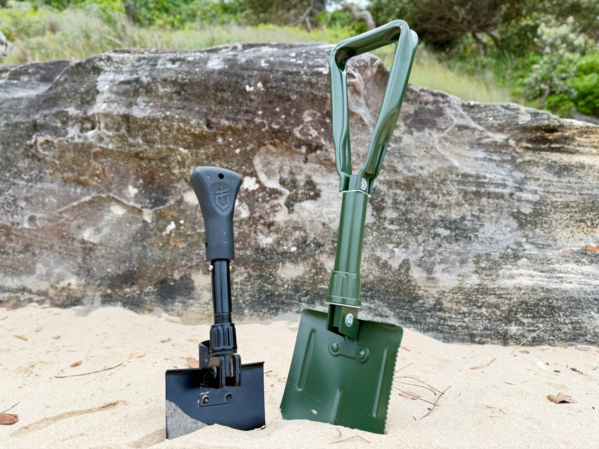 The green OZtrail Tri-Fold Shovel standing in the sand next to the smaller black Gerber Gorge