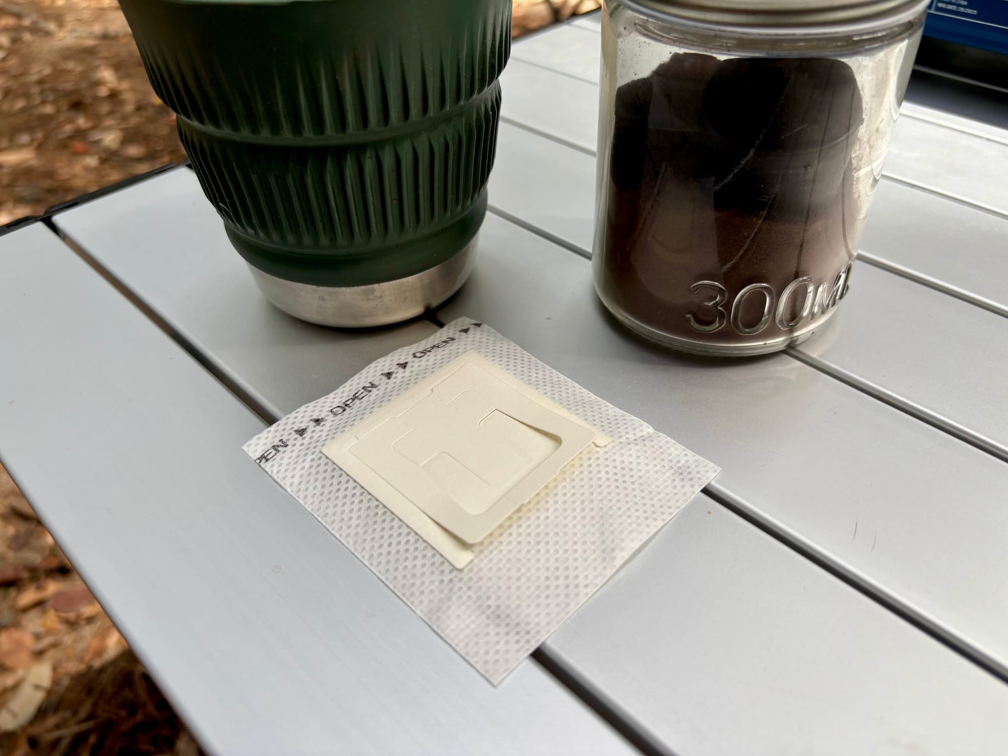 A single drip coffee filter bag lying flat, with a mug and a jar of ground coffee