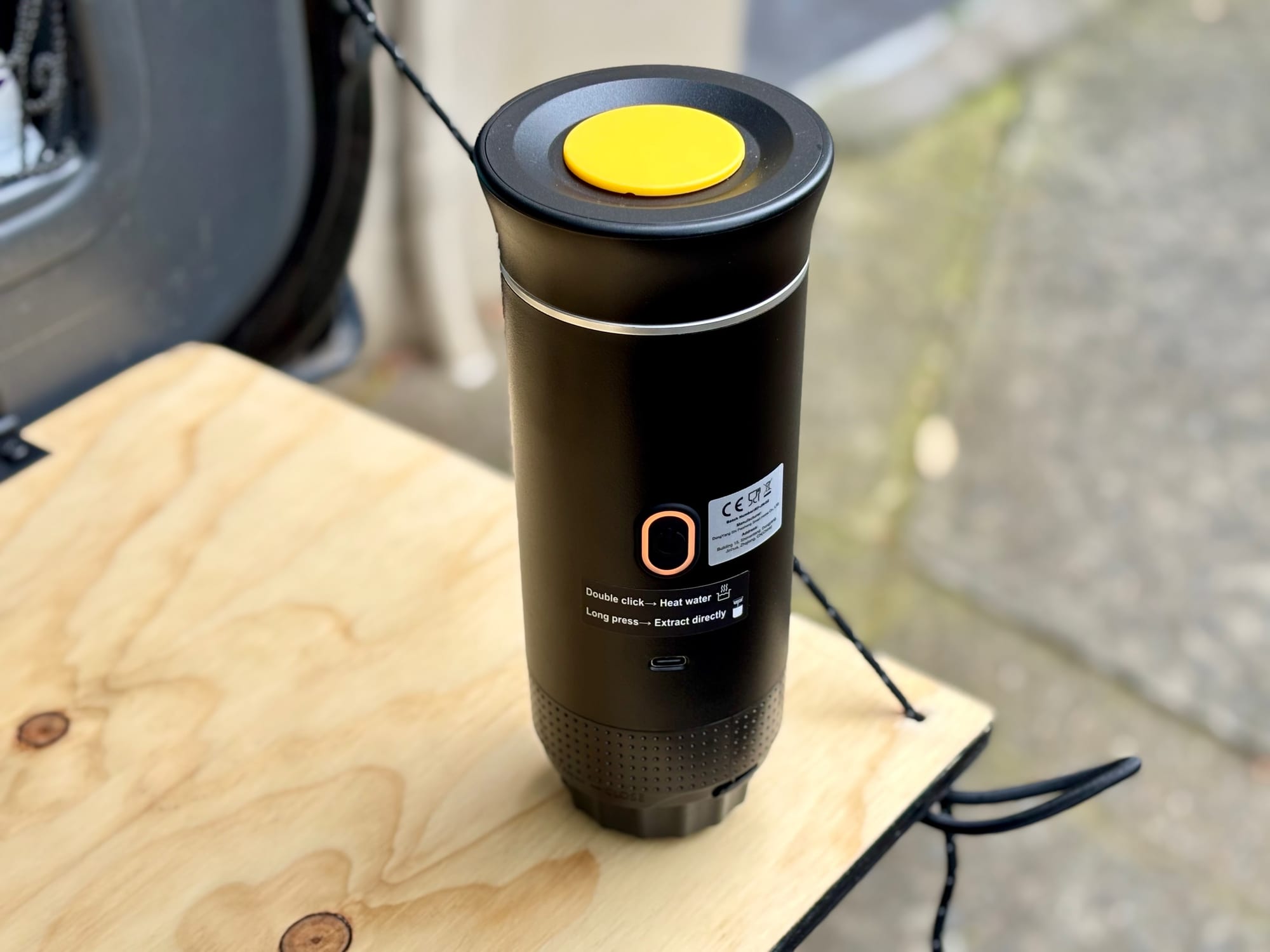 The portable electric espresso machine with its yellow lid on, standing on a small camp table