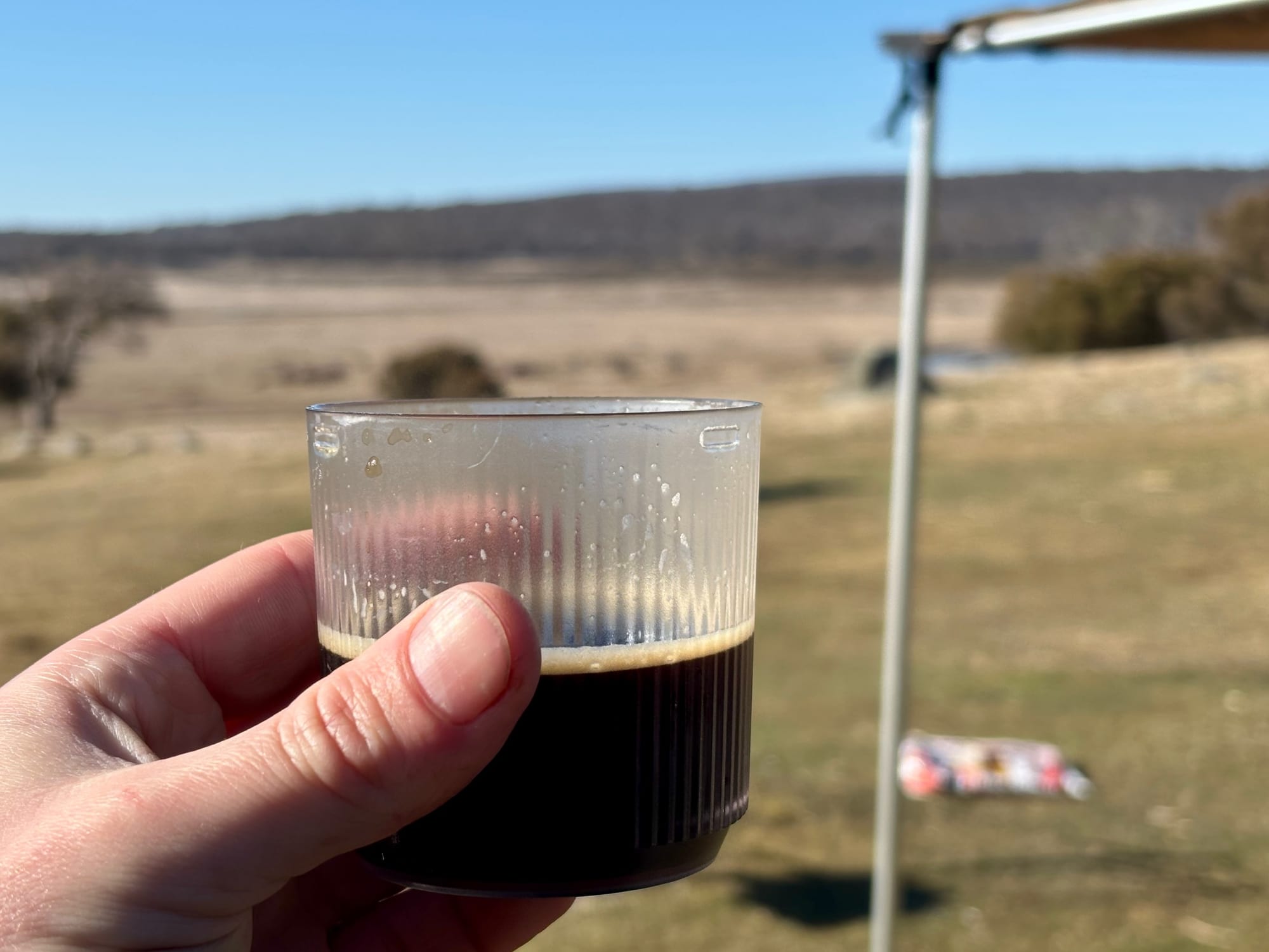 A hand holding a clear cup of espresso at an open campsite, with grassy plains behind