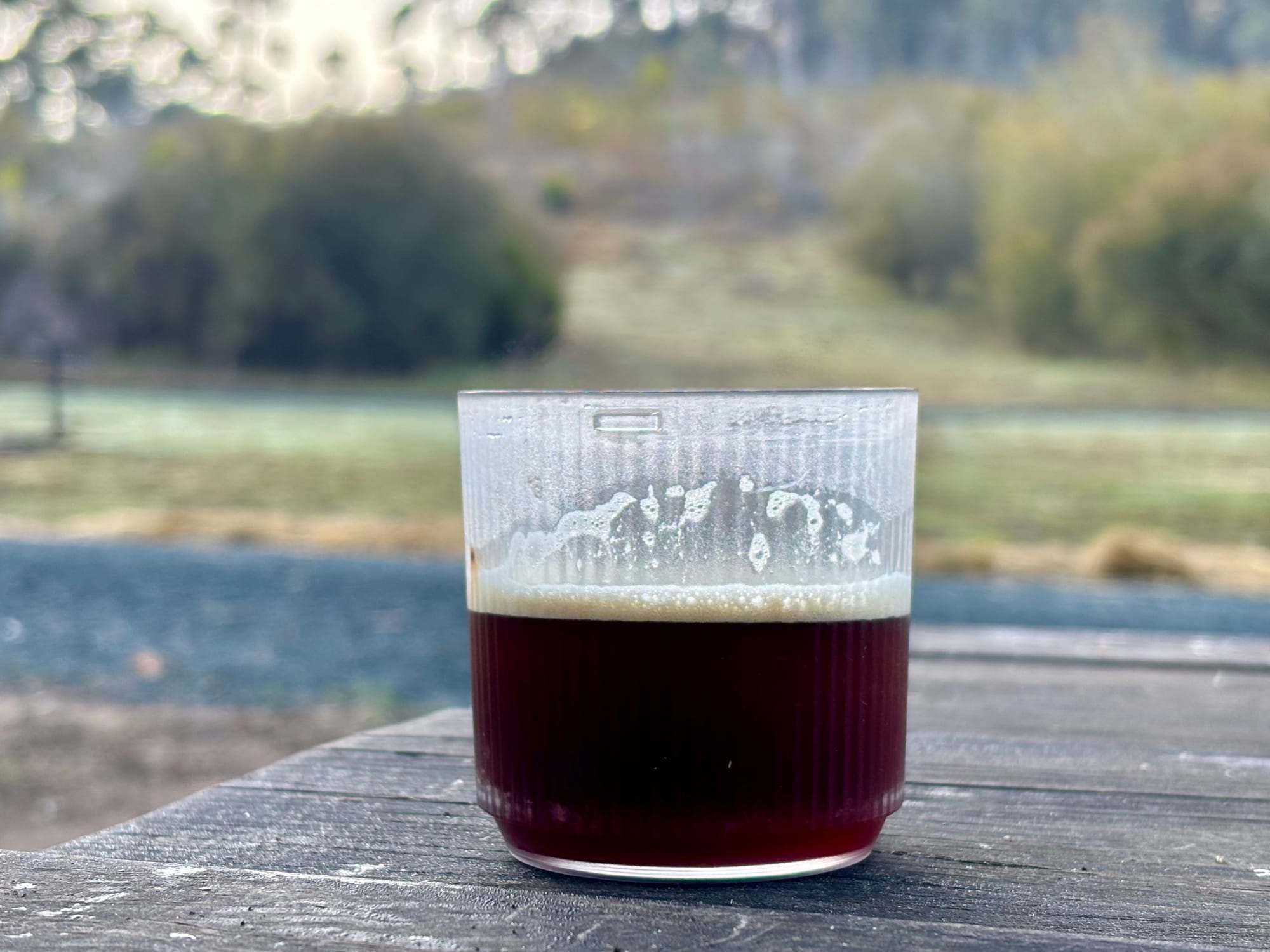 A clear cup of espresso with a layer of crema, standing on a picnic table at camp