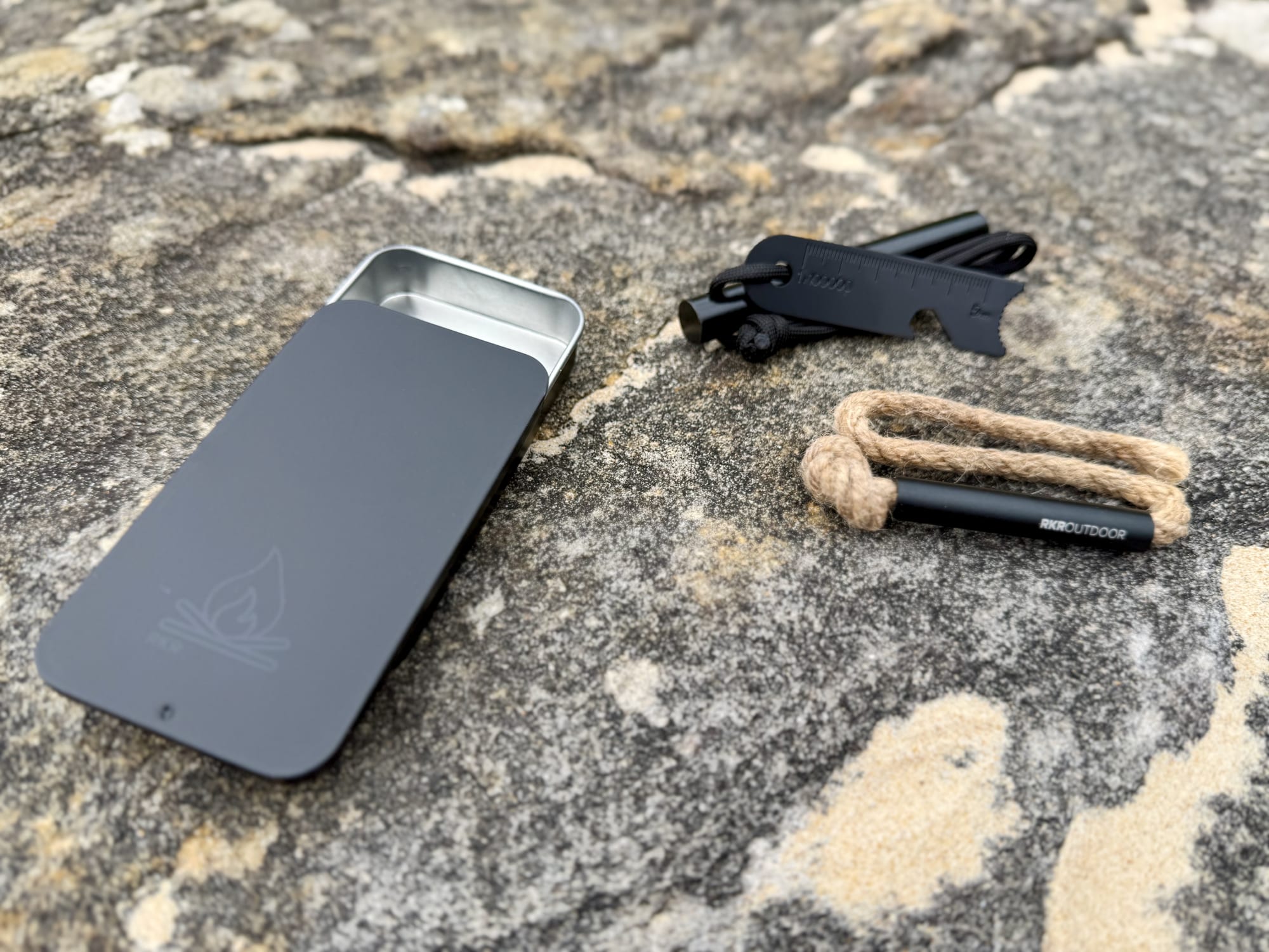 The tin, ferro rod with striker and tinder wick of the RKR Outdoor kit next to each other