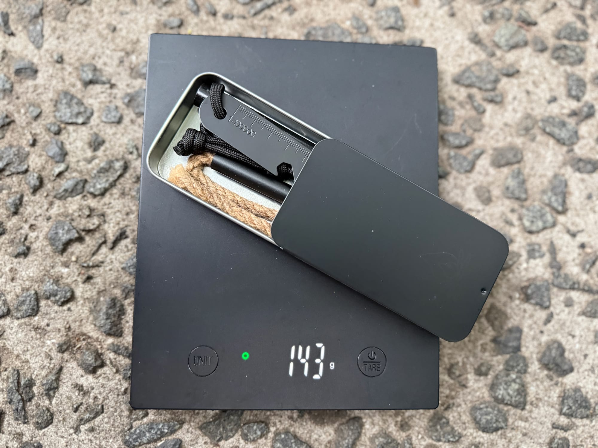 The RKR Outdoor fire starter kit in its tin on a scale showing 143 g
