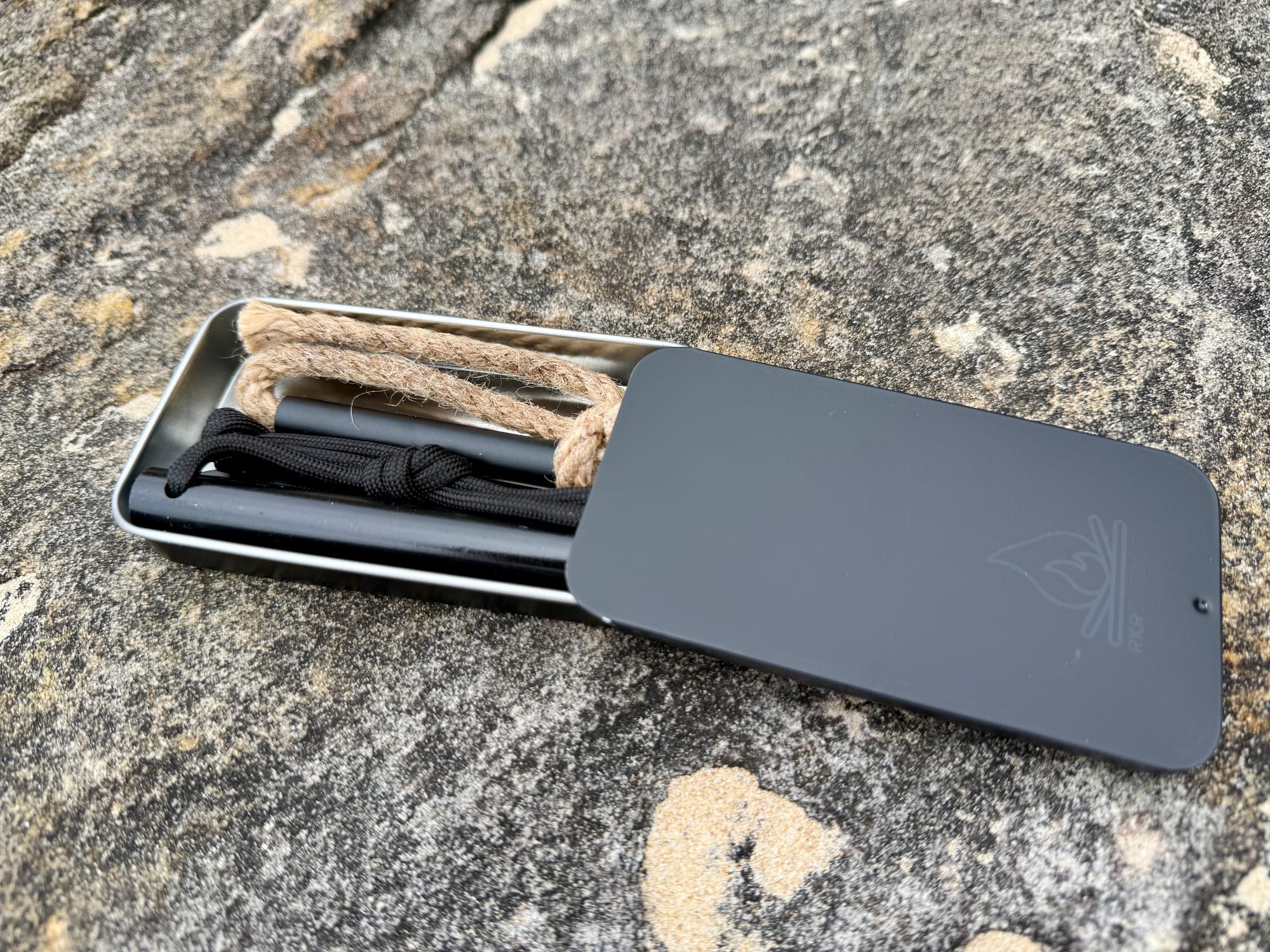 The open tin of the RKR Outdoor fire starter kit with the rod, striker and tinder wick inside