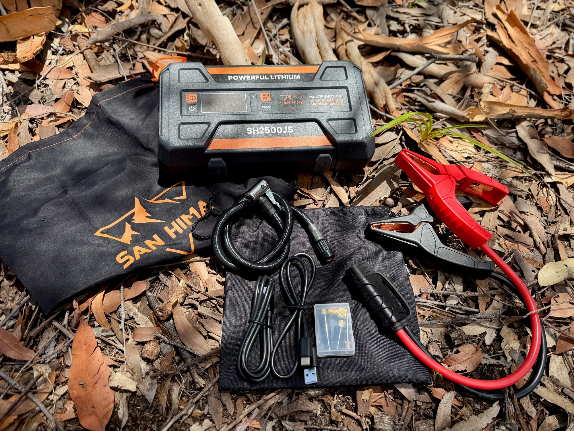 The San Hima jump starter with its storage bag, the red and black jump leads, the air hose, cables and manual, laid out on leaf litter