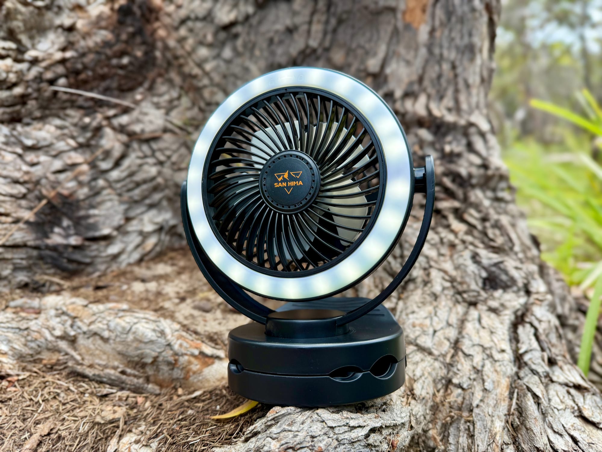 The San Hima clip fan standing on its base on tree roots, with the LED ring light on