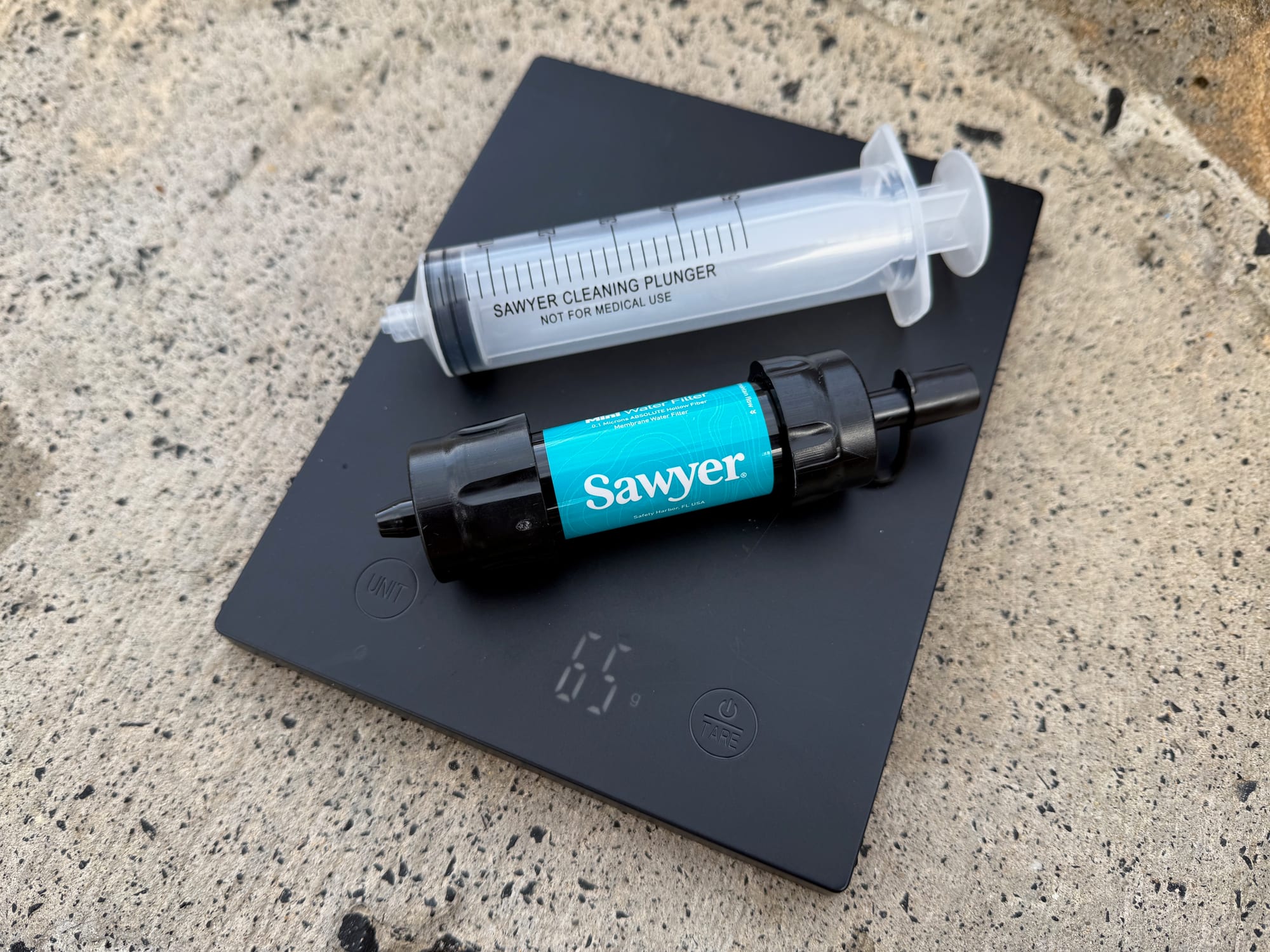 The Sawyer Mini filter and its cleaning plunger on a kitchen scale showing 65 g