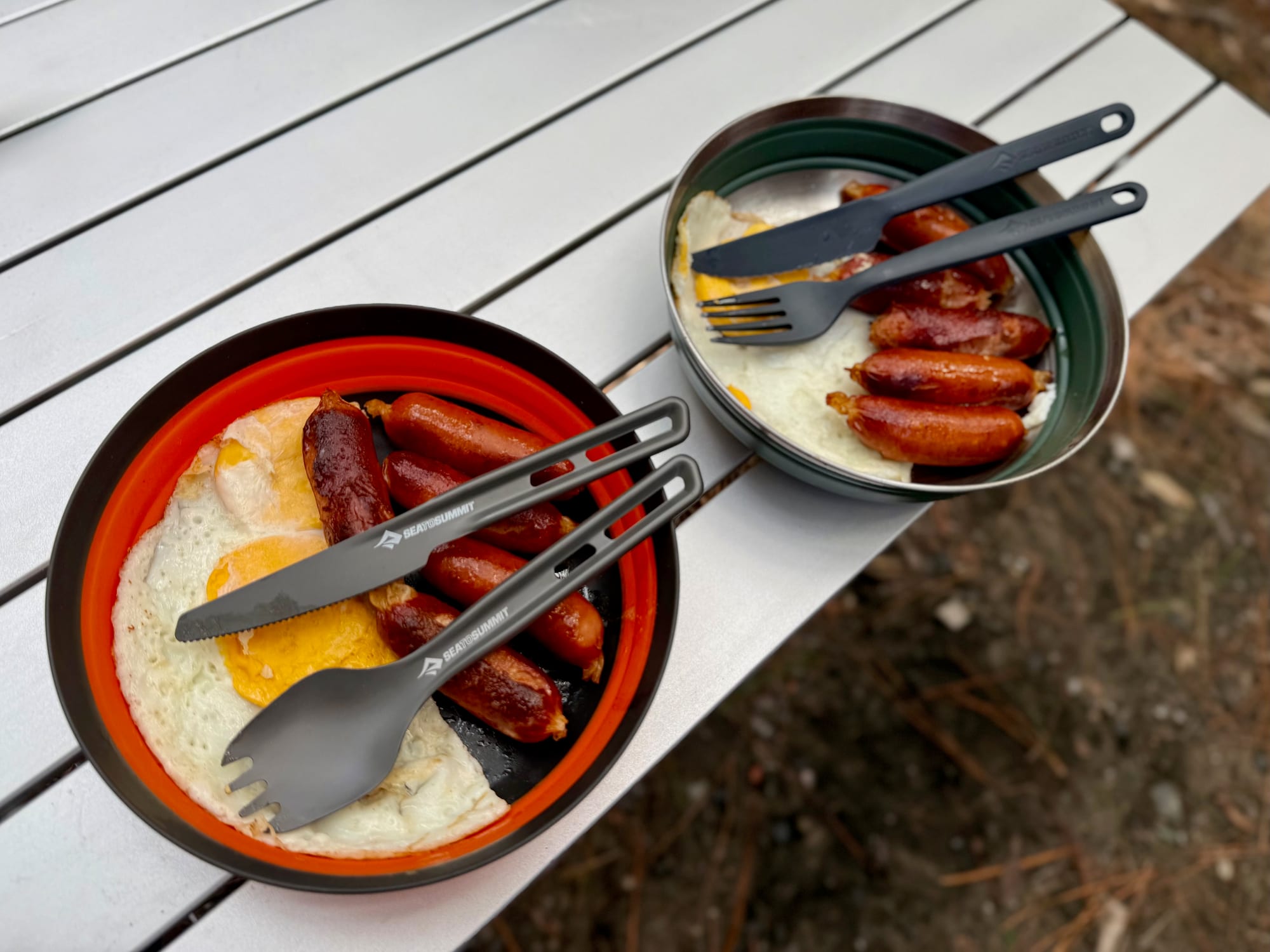 A green Detour bowl with sausages next to an orange Frontier bowl with eggs and sausages on a camp table