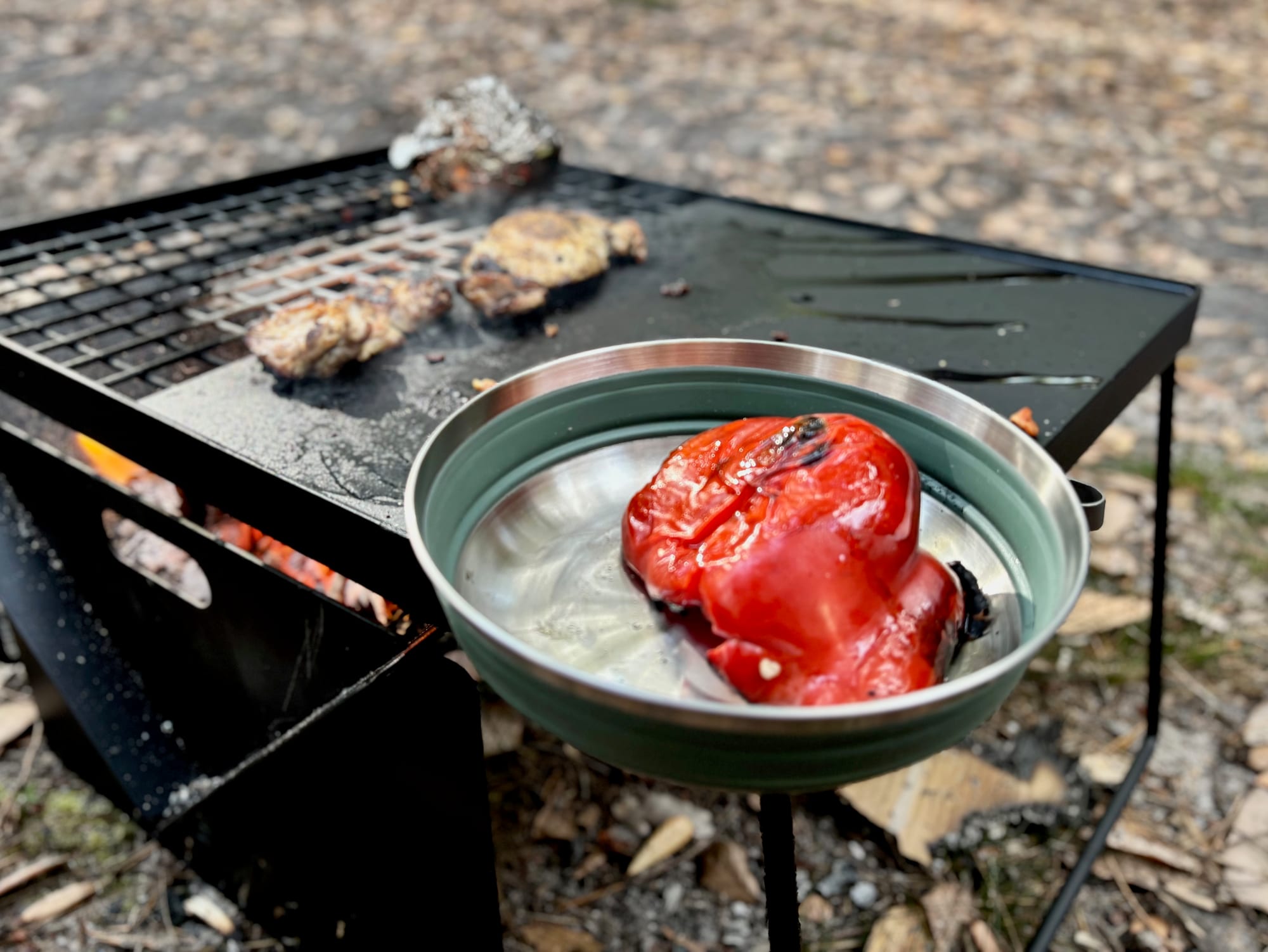 A green Detour collapsible bowl with a grilled red capsicum, on a fire grill next to sausages