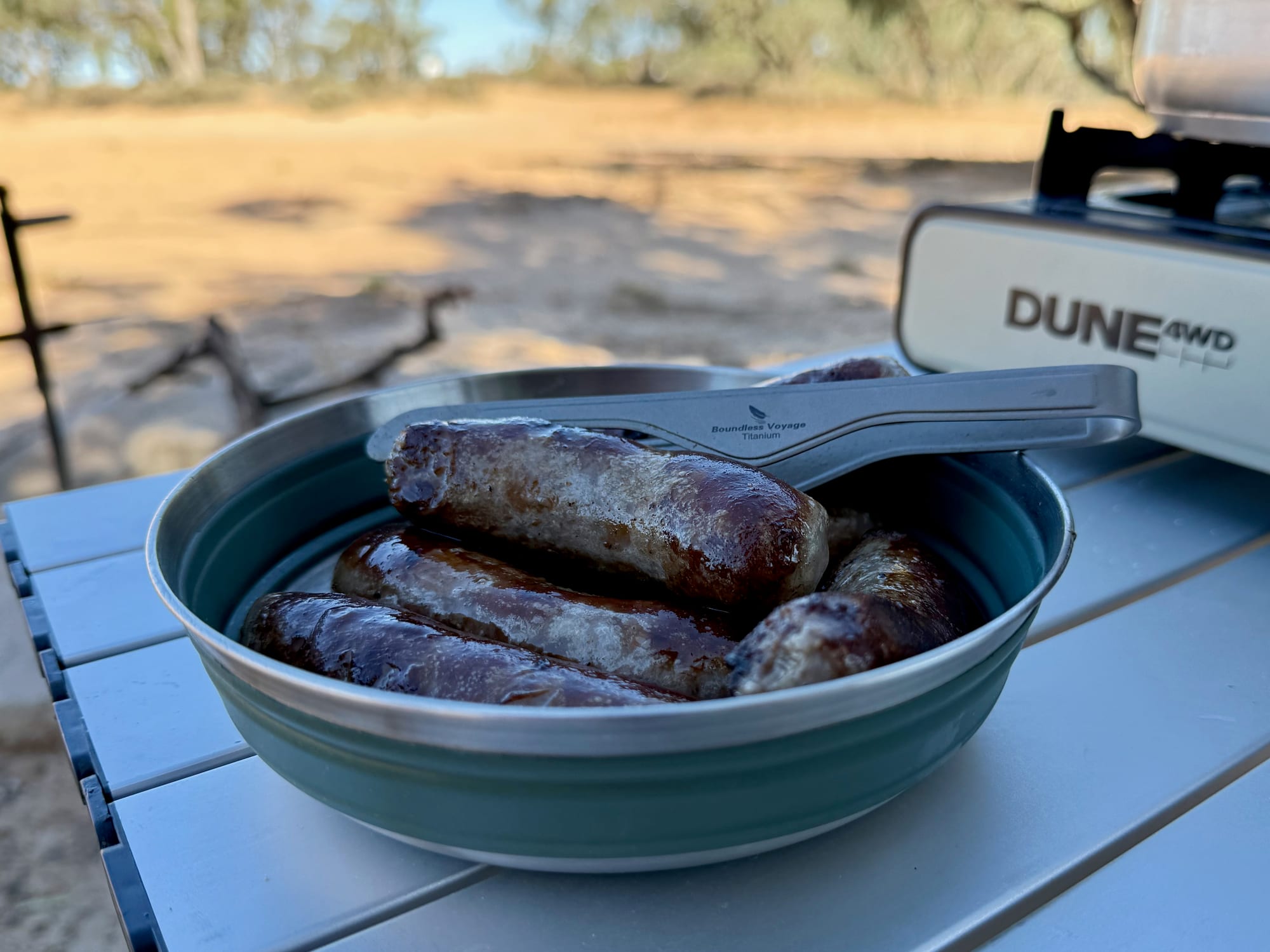 A green Detour bowl with grilled sausages and a spork on a camp table, a Dune stove behind it