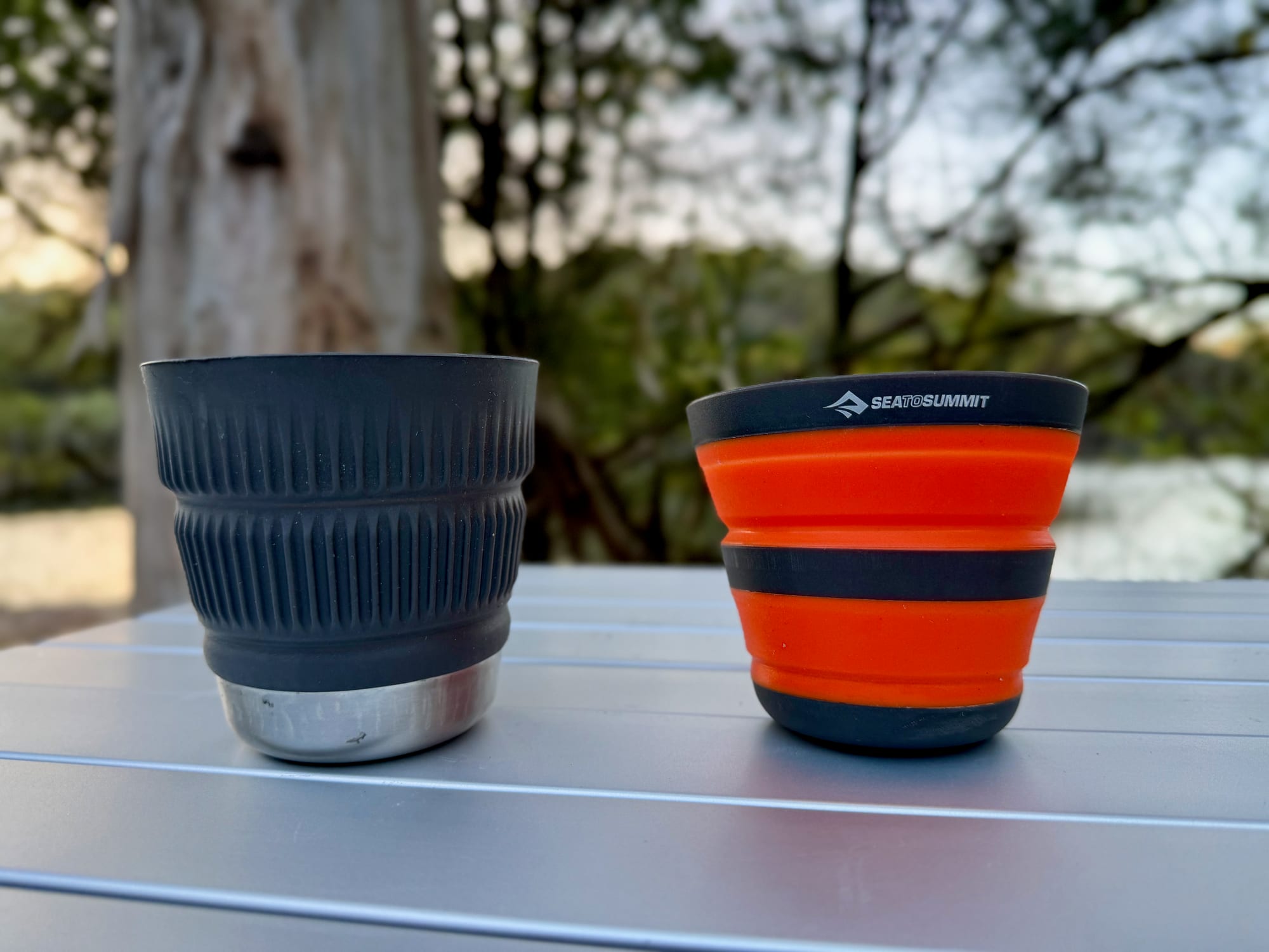 The Beluga Detour mug and the orange Frontier collapsible cup open side by side on a camp table