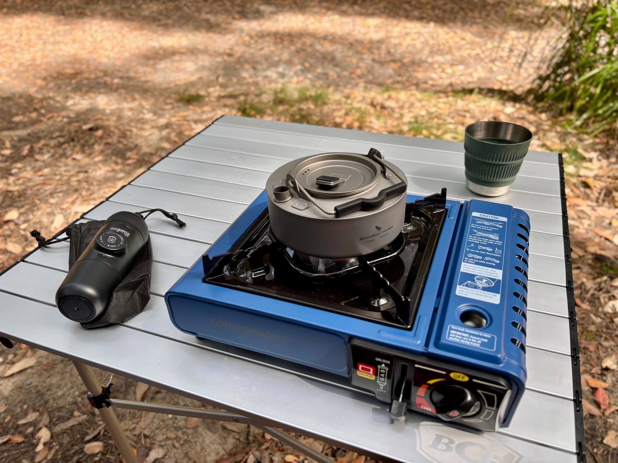 The green Detour mug on a camp table next to the titanium kettle on a gas stove