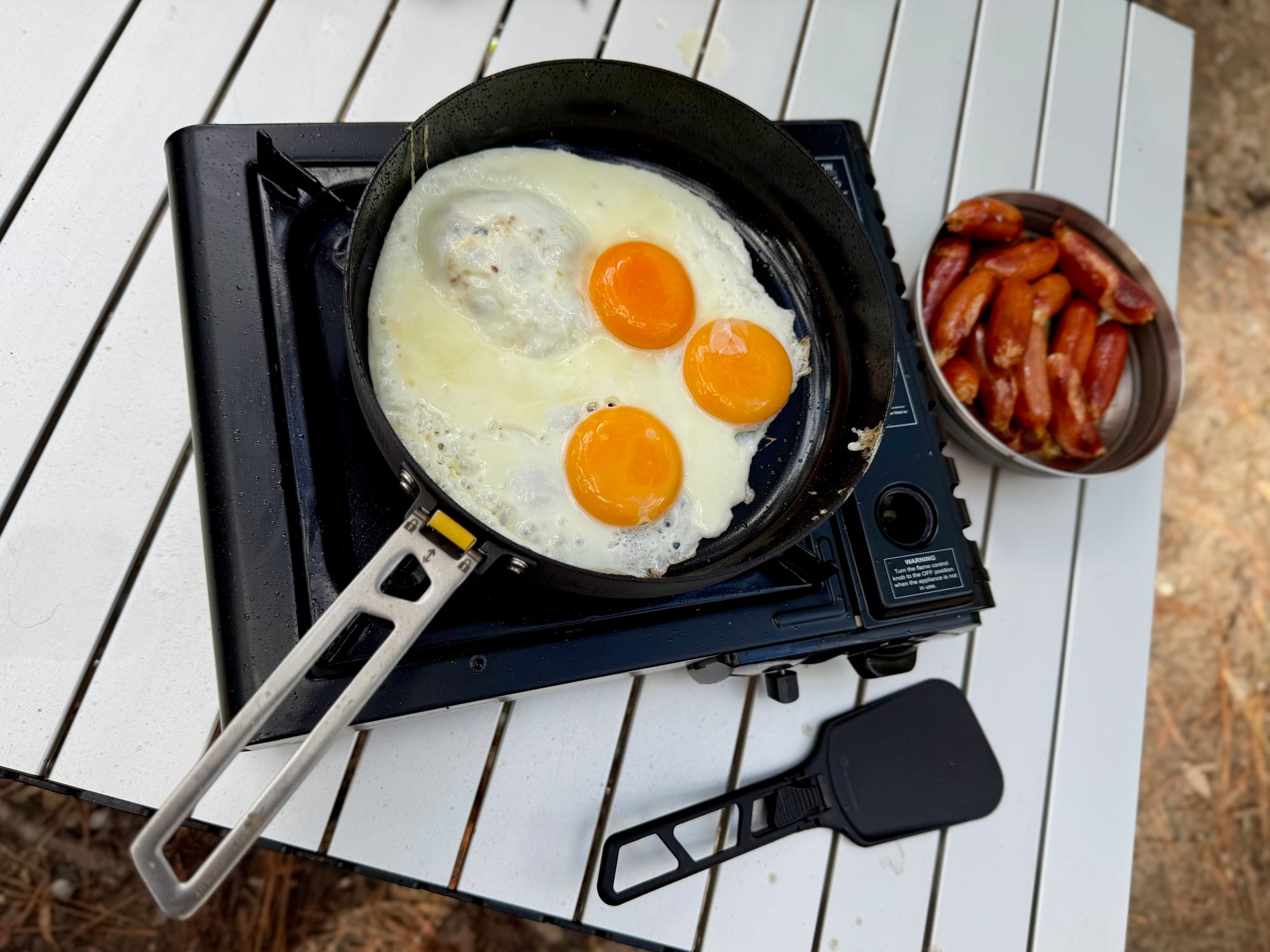 Three fried eggs in the Sea to Summit Frontier Ultralight Pan on a butane stove, with a bowl of sausages beside it on a white camp table