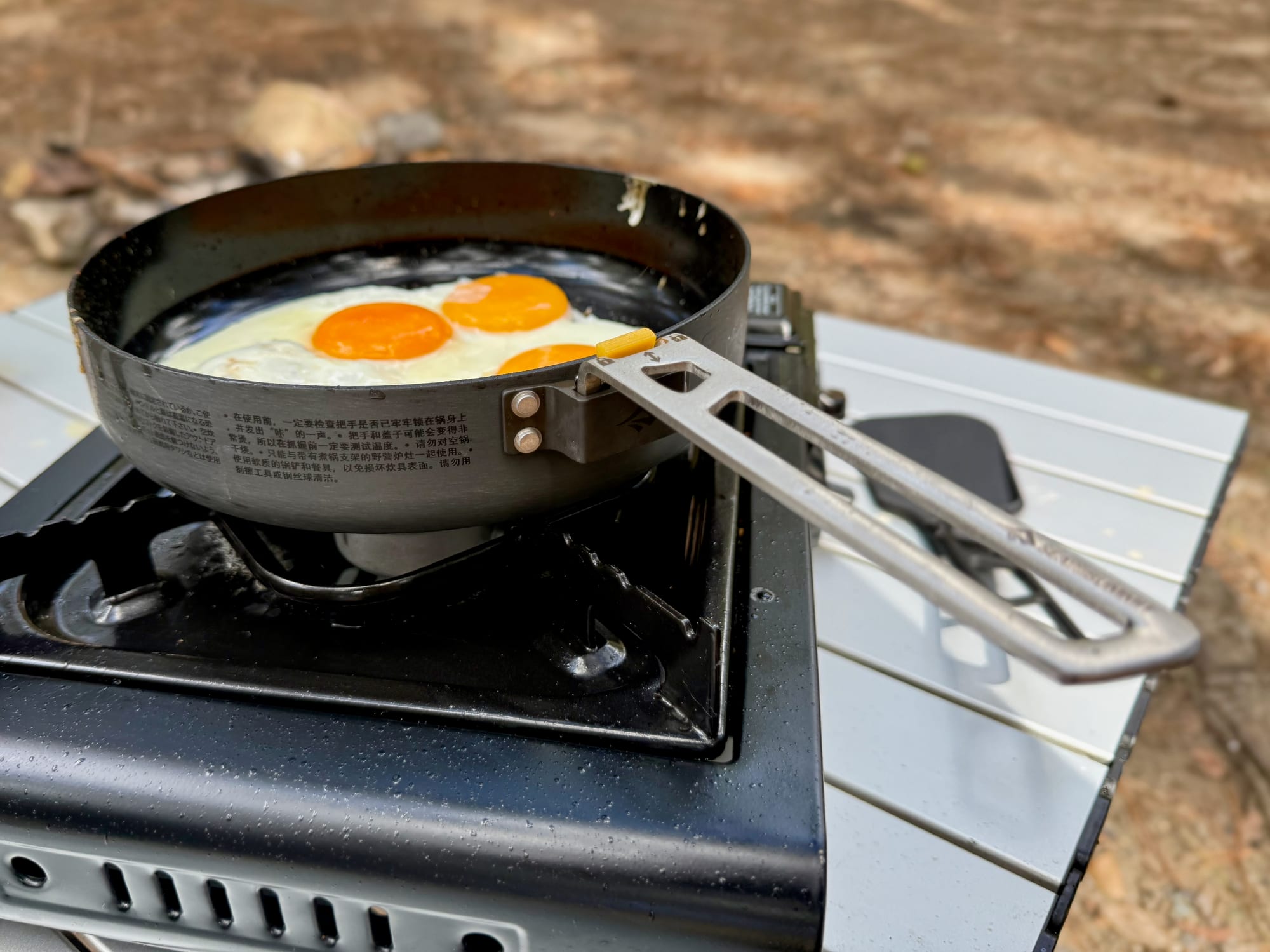 Eggs frying in the Frontier Ultralight Pan on a butane stove, with its long stainless steel handle clicked on and reaching over the camp table