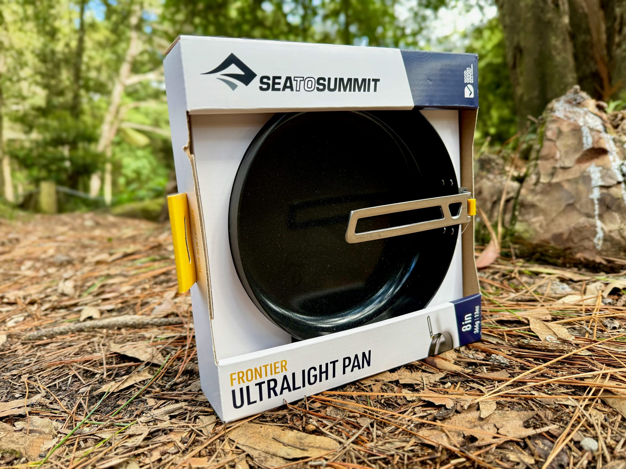 The Sea to Summit Frontier Ultralight Pan in its box, with the handle showing, on pine needles under the trees