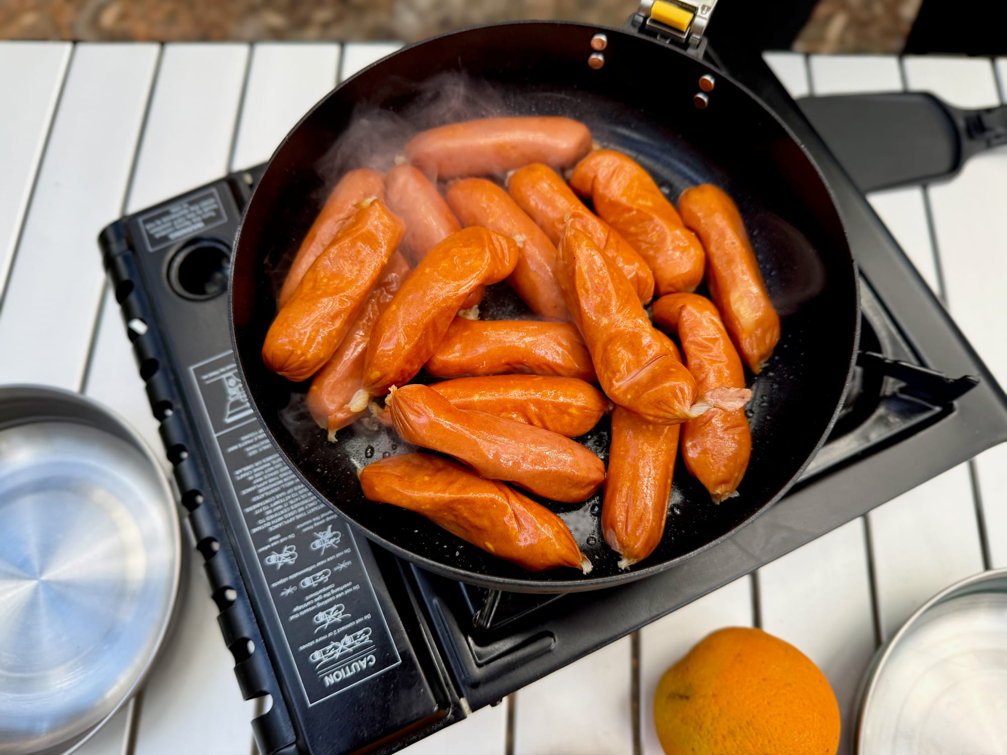 A panful of sausages in the Frontier Ultralight Pan from above, on a butane stove on a white camp table