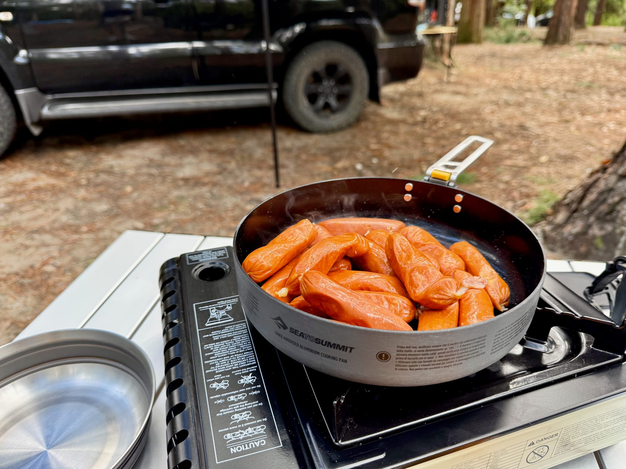 Sausages in the Frontier Ultralight Pan on a butane stove on a camp table, with a stainless steel plate beside it and the Prado behind