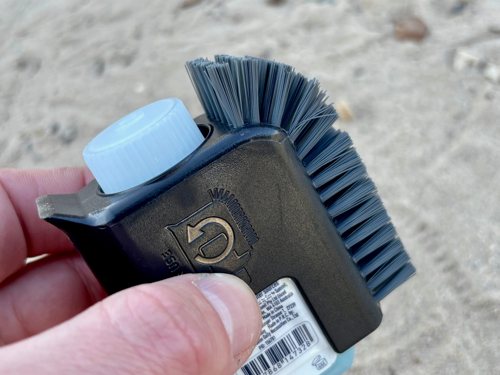 The Sea to Summit Pot Scrubber held in one hand, showing the scraper edge and the bottle clipped in