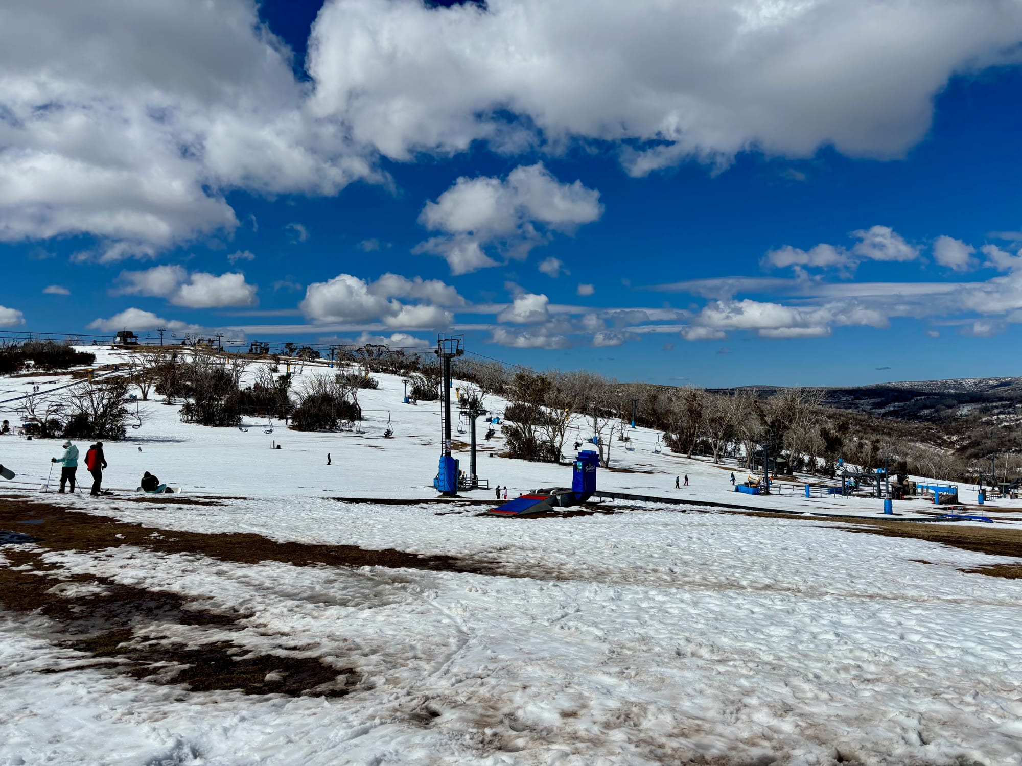 The slopes of Selwyn Snow Resort with skiers, a chairlift and patchy late-season snow under a blue sky