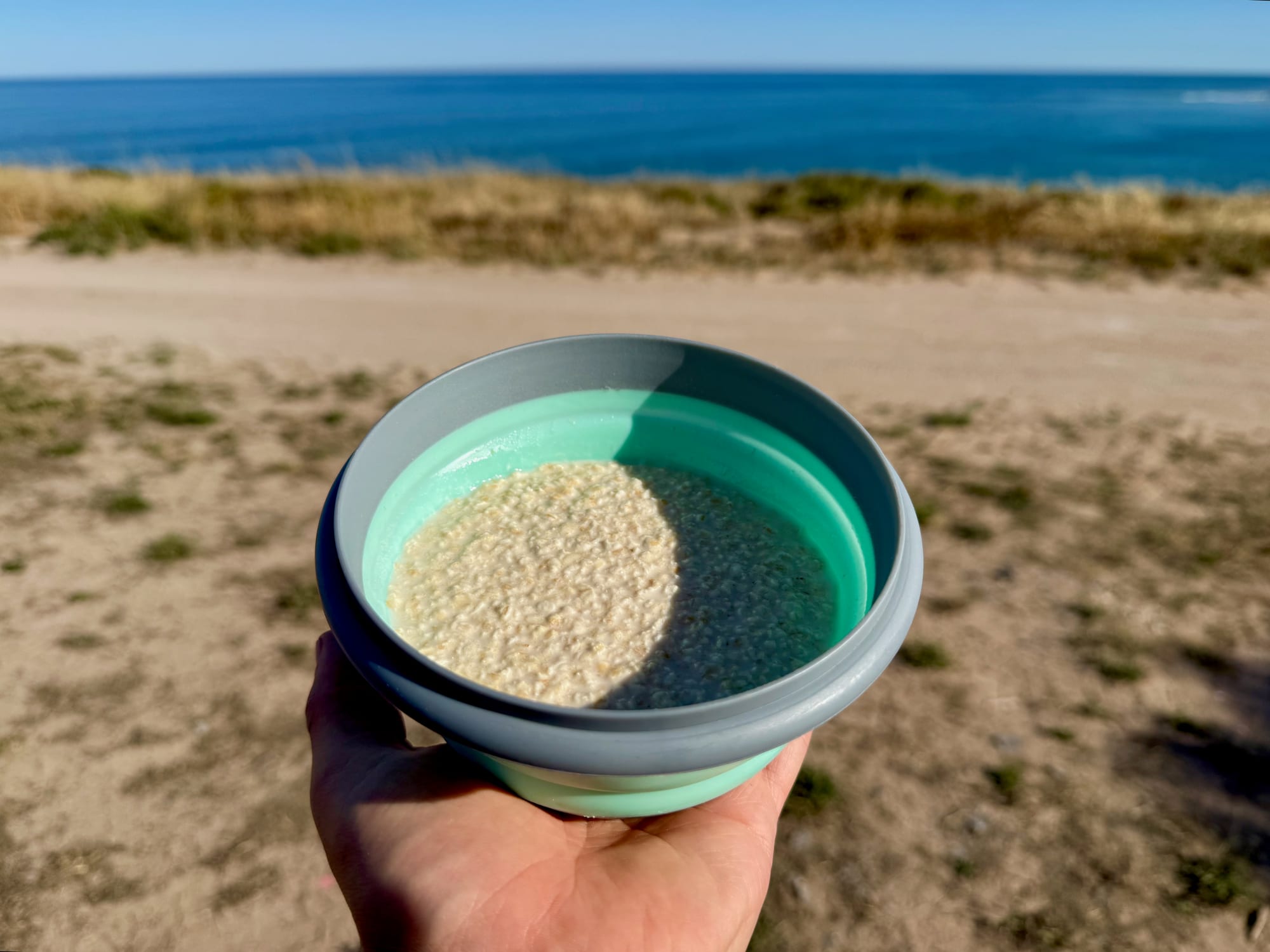 A teal silicone collapsible bowl full of rice, held up by the sea