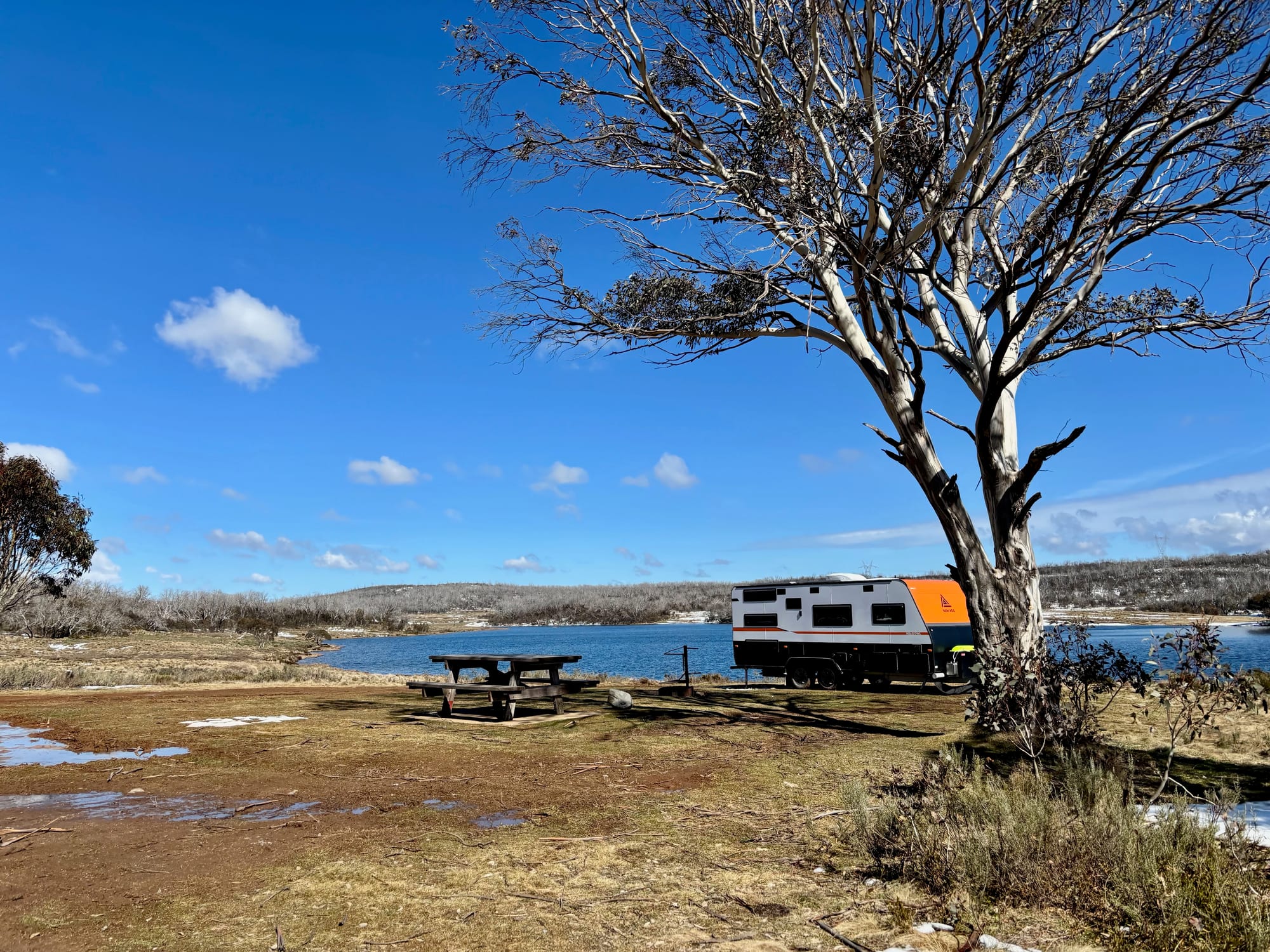 A picnic table and a caravan under a snow gum on the shore of Three Mile Dam