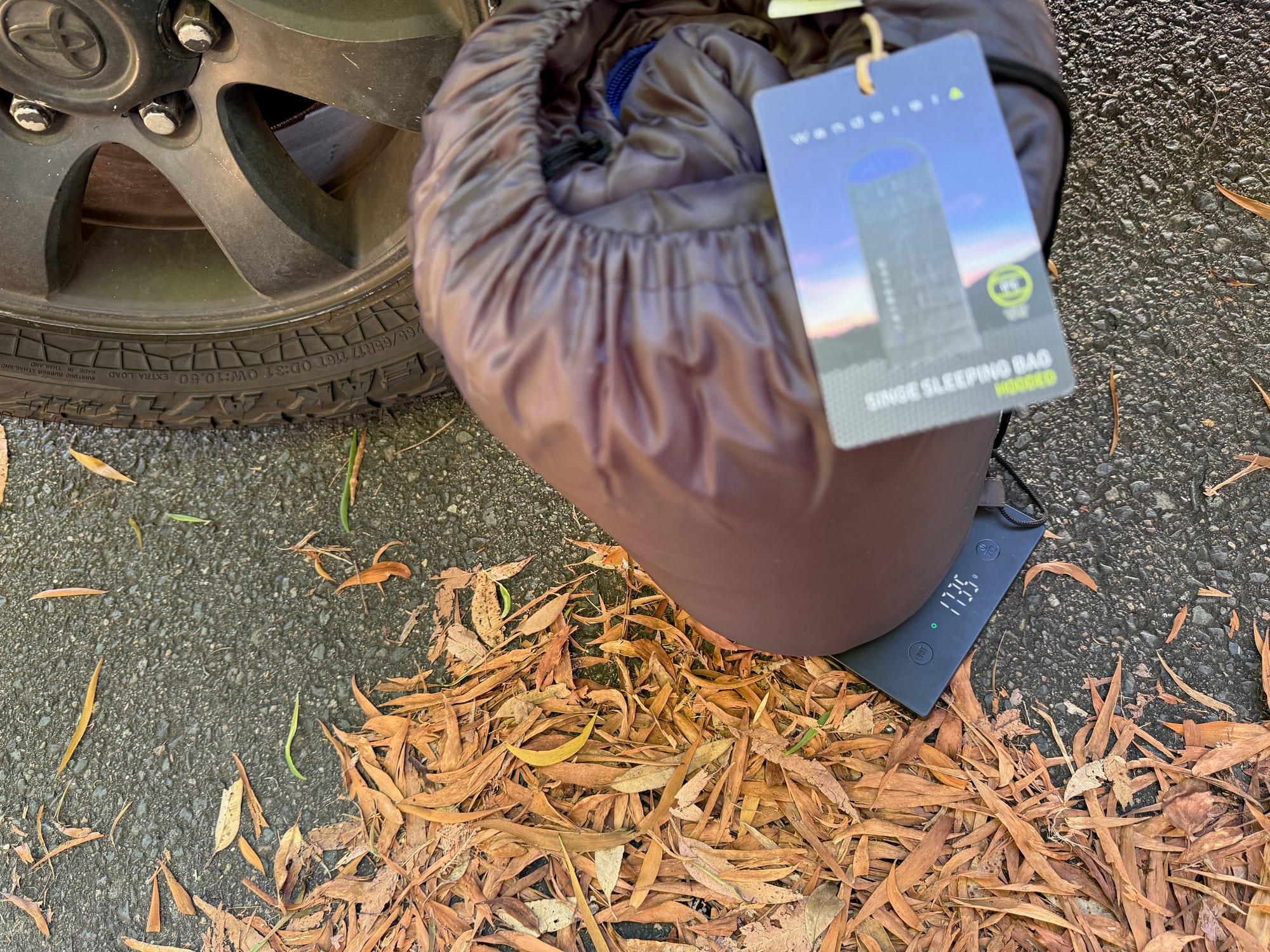 The Wanderer sleeping bag in its stuff sack with the swing tag, on a scale next to the Prado’s wheel