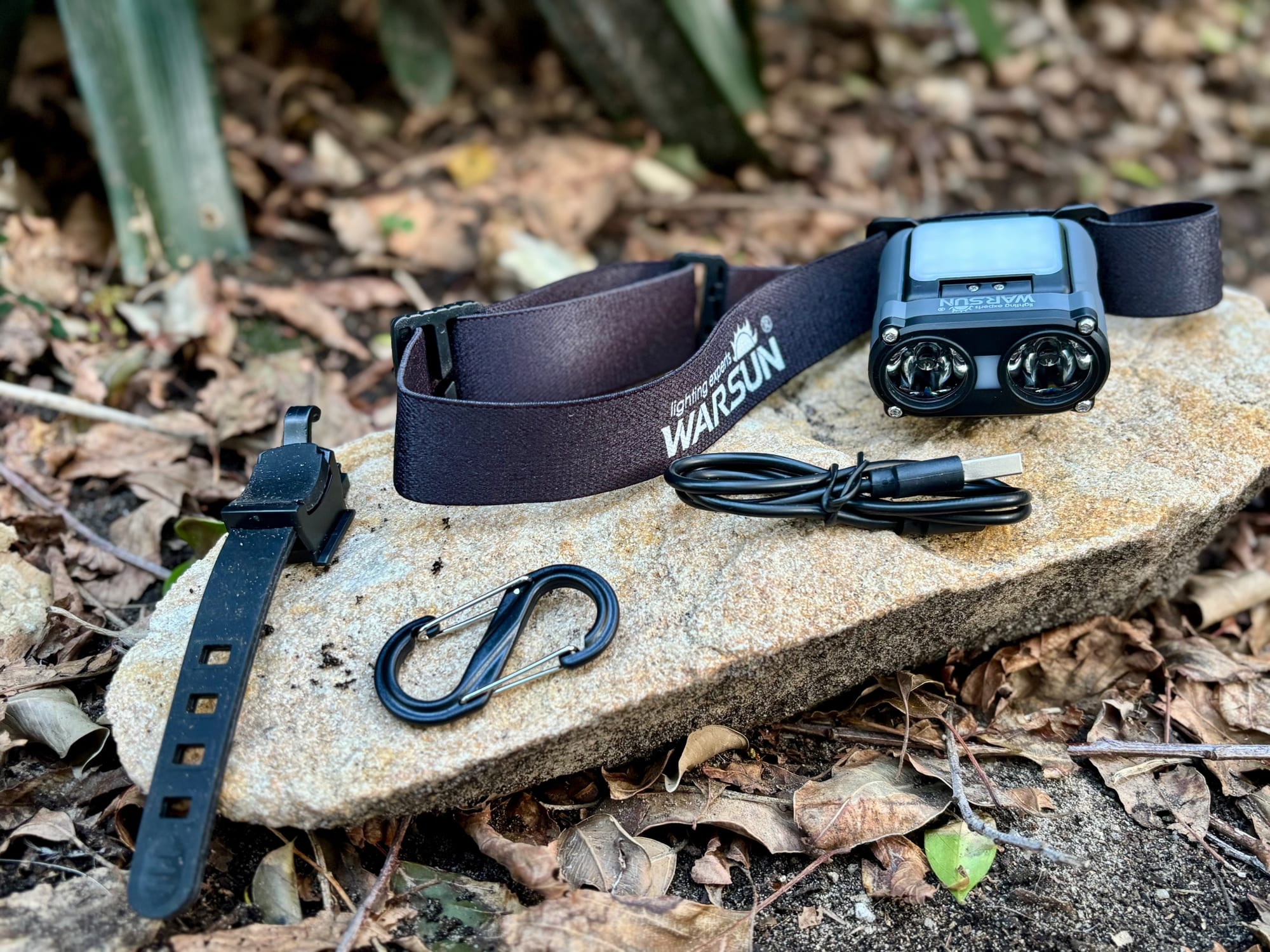 The Warsun headlamp with its headband, bike strap, charging cable and a small carabiner laid out on a rock