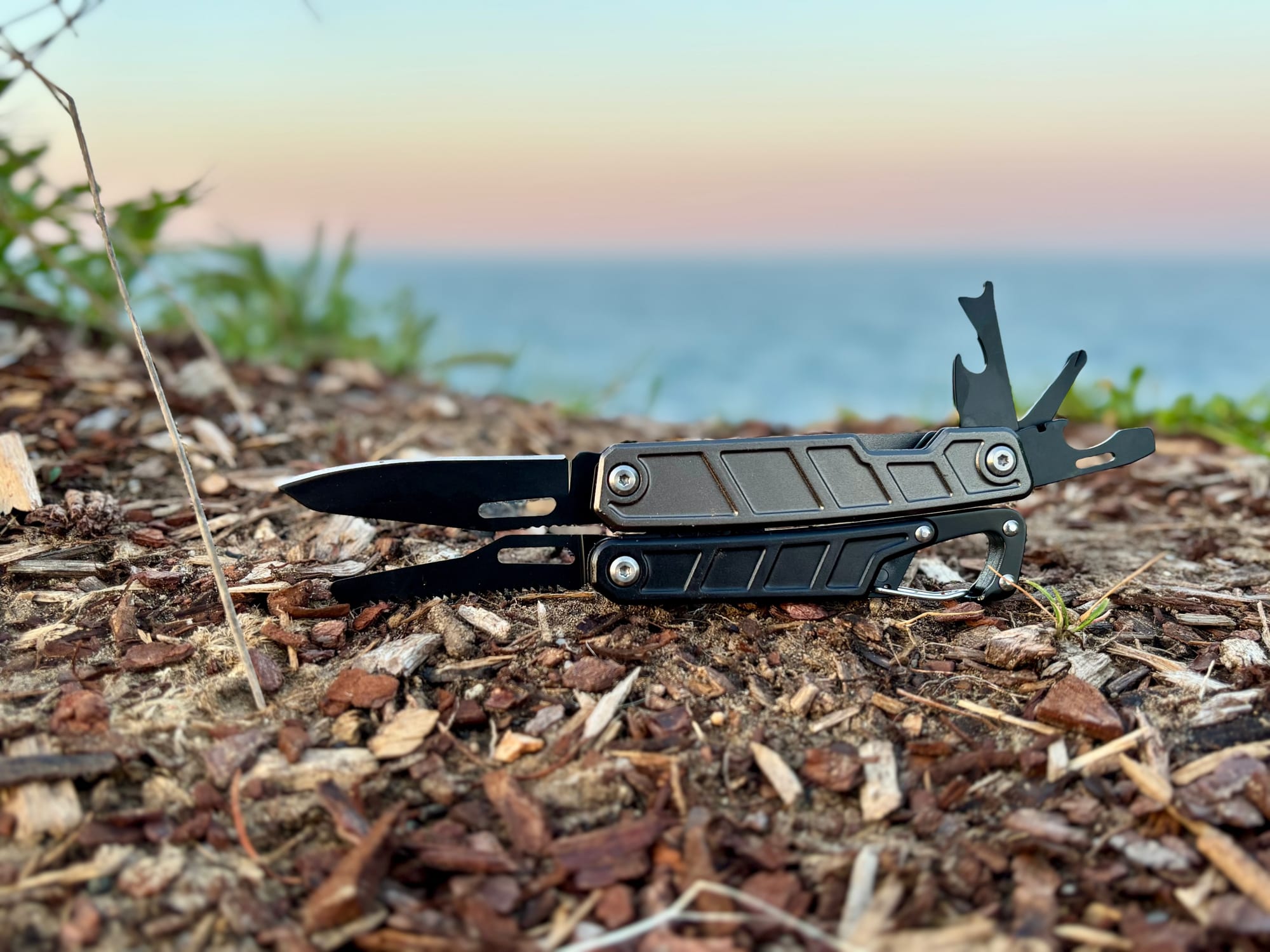 The Workpro 11-in-1 multitool with its knife, saw and small tools all folded out, with the sea and a sunset sky behind