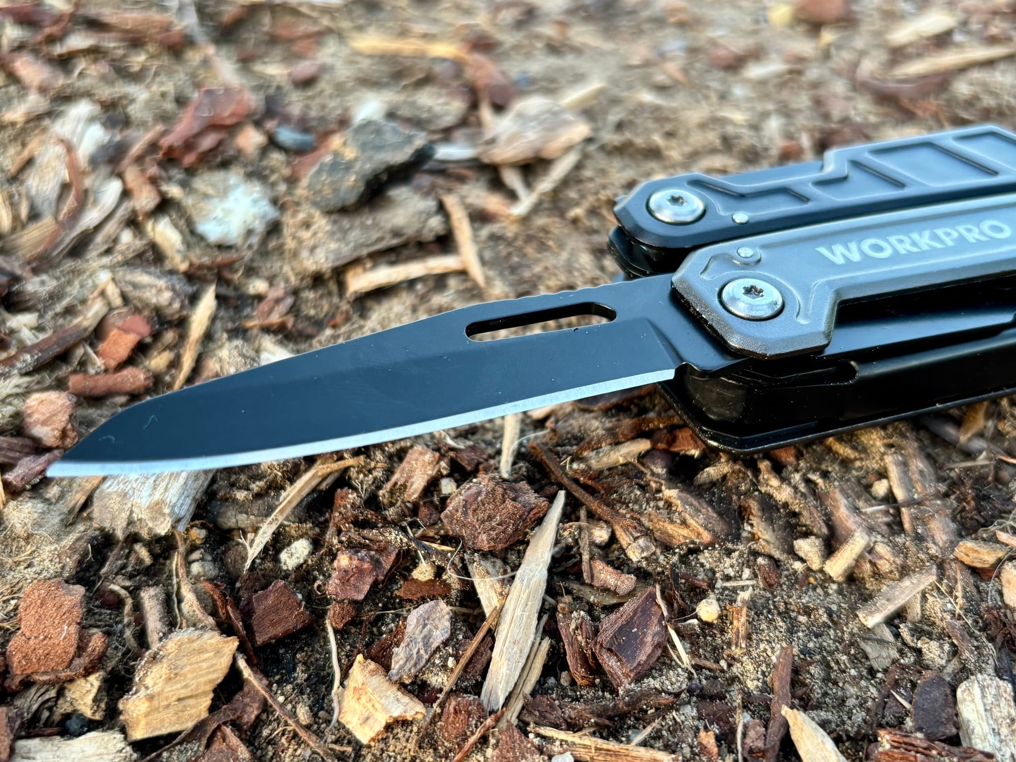 The black knife blade of the Workpro 11-in-1 multitool folded out, lying on wood chips