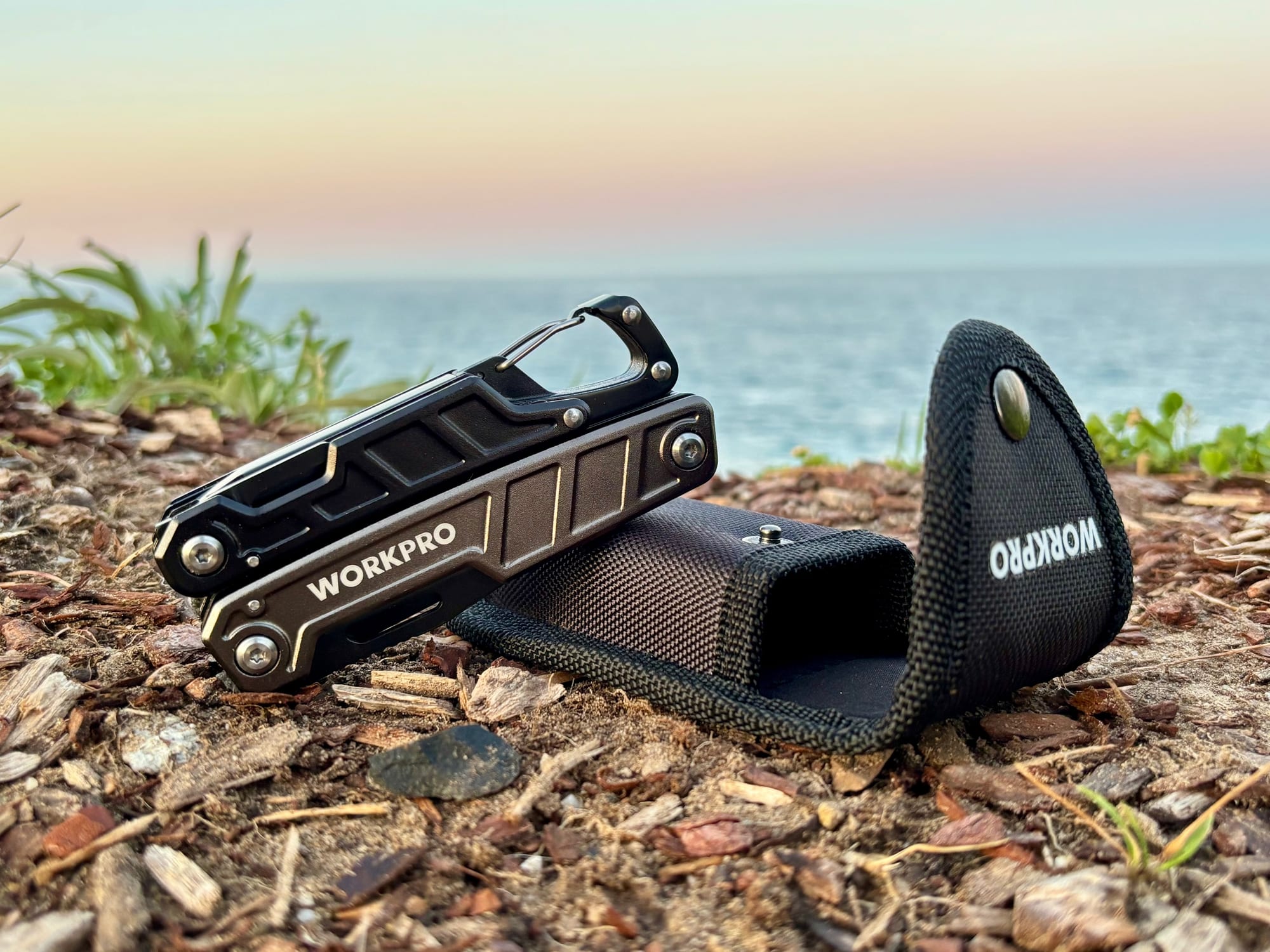 The Workpro 11-in-1 multitool leaning on its open nylon sheath, with the sea behind