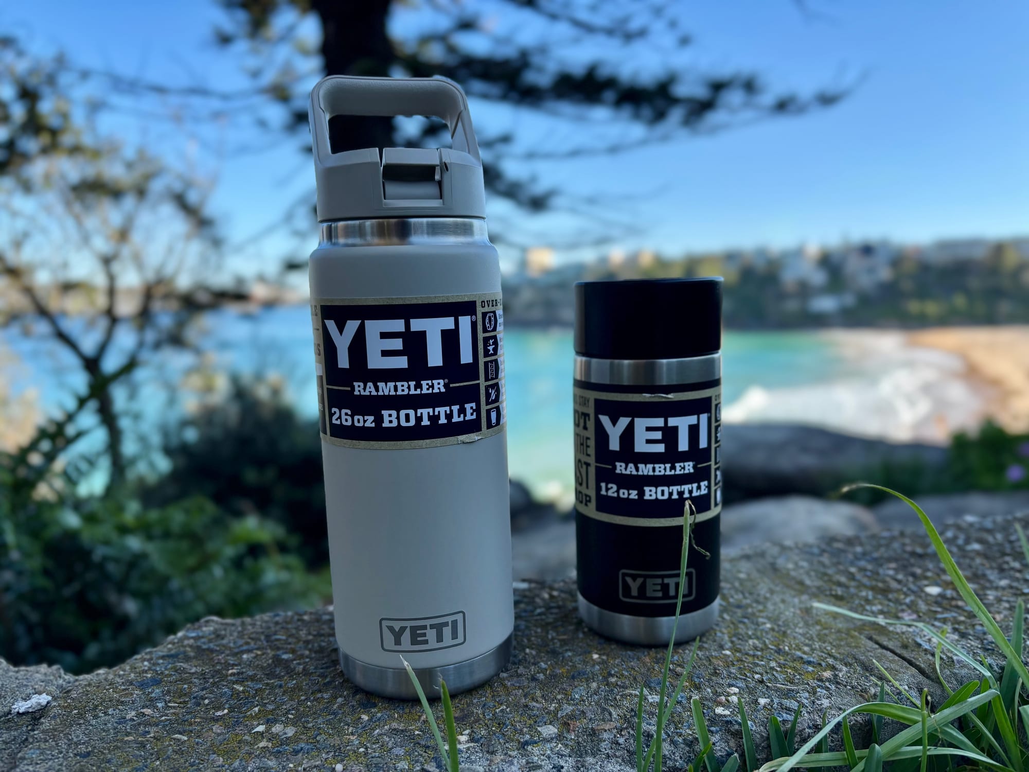 The black YETI Rambler 12 oz bottle next to the taller Cape Taupe 26 oz bottle, with a beach behind