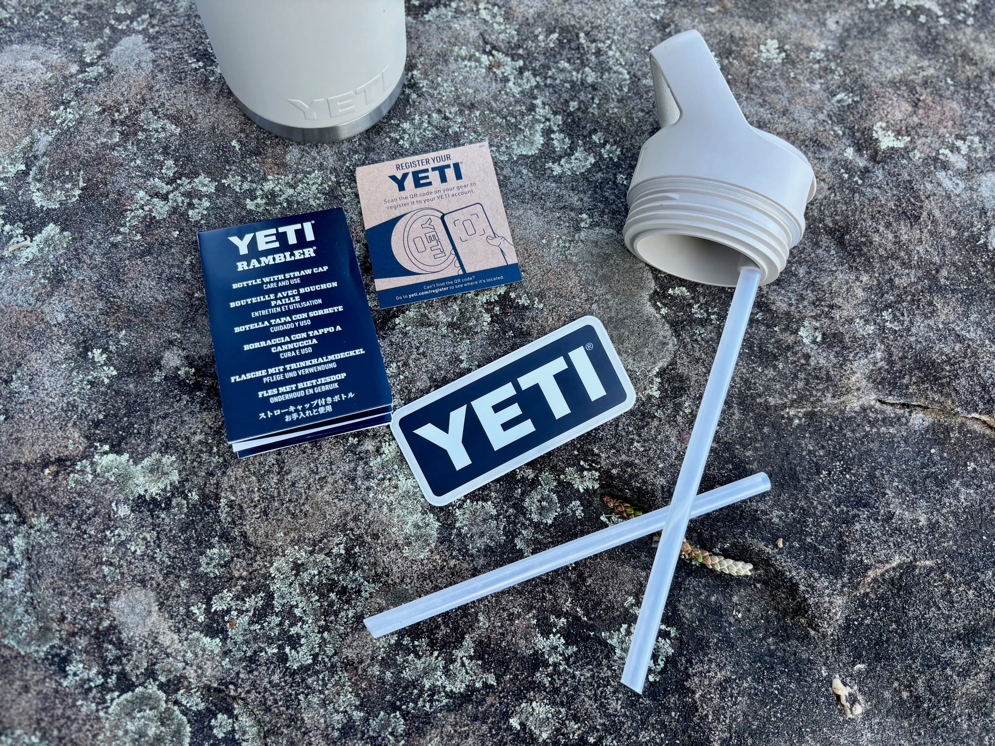 The straw cap with its straw, a spare straw, the care leaflet and a YETI sticker laid out on a rock