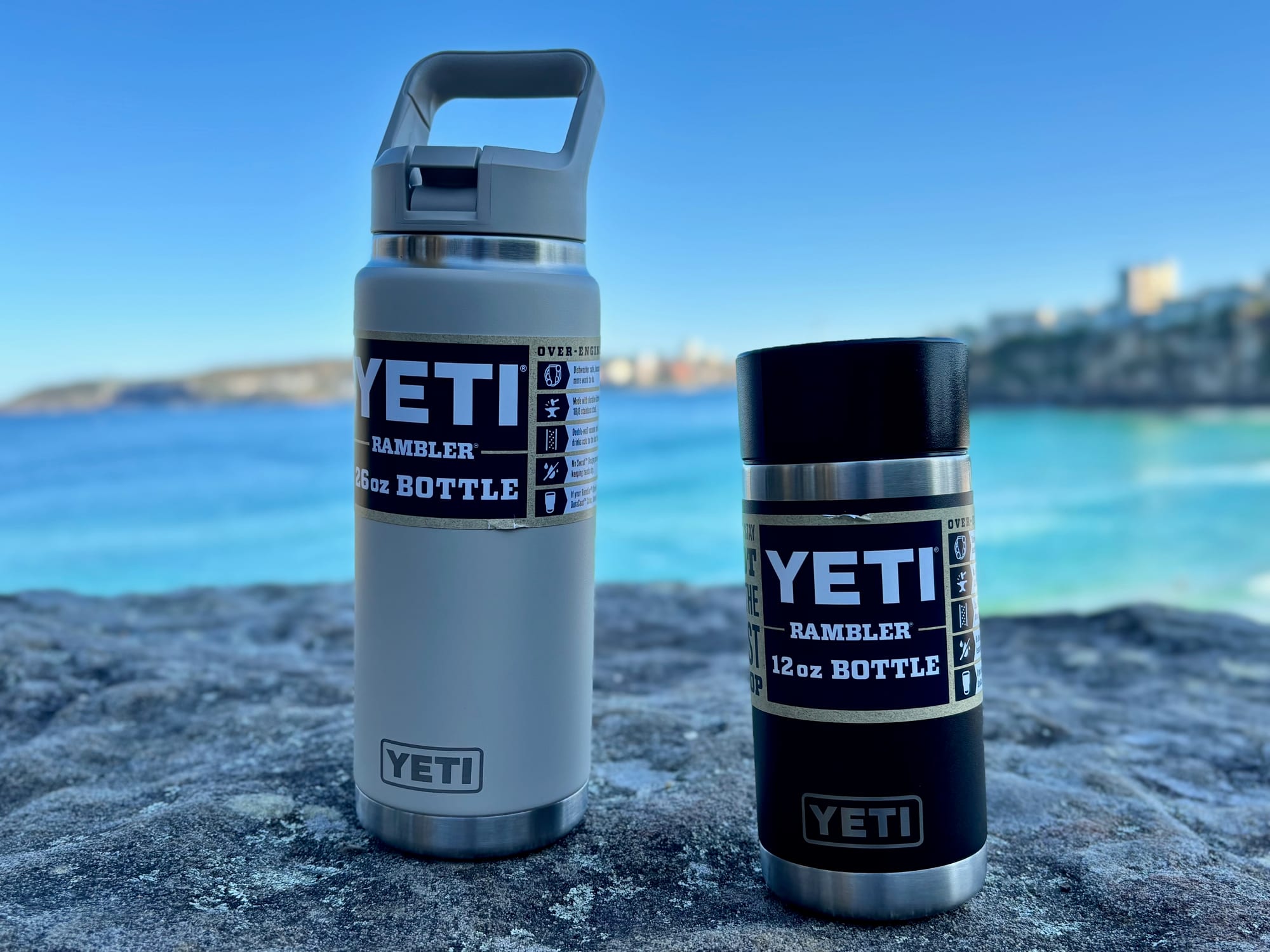 The Cape Taupe YETI Rambler 26 oz bottle next to the smaller black Rambler 12 oz bottle on a rock above the sea