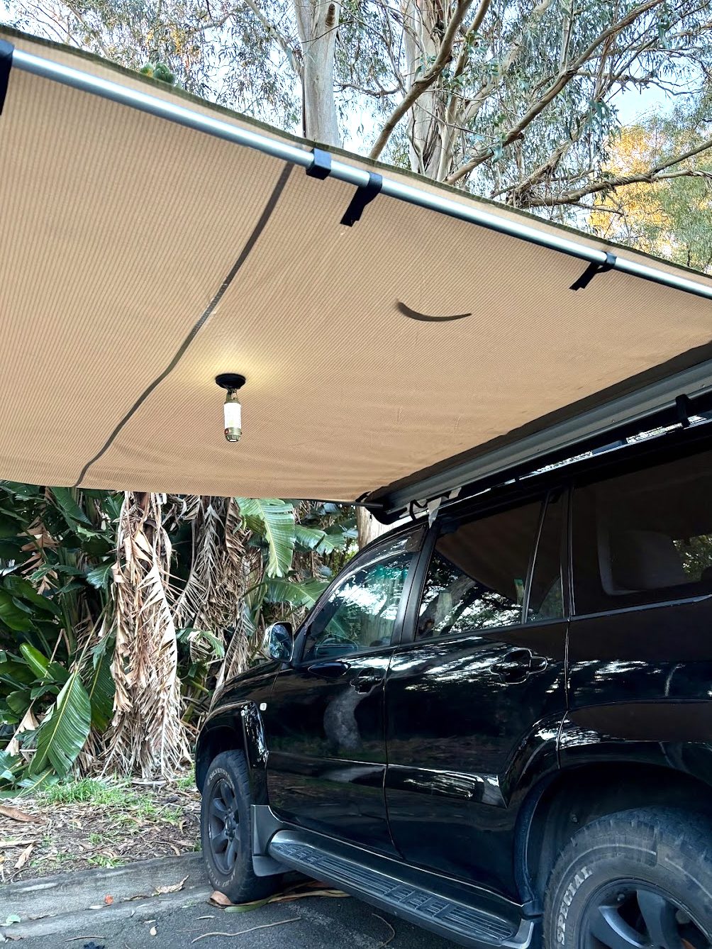A camp light hanging from a magnetic hook under the awning, with the car and trees behind